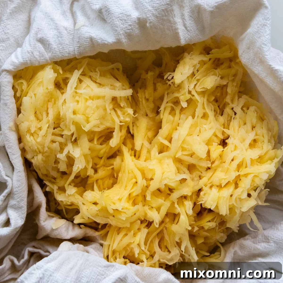 Close-up of raw grated Yukon gold potatoes bundled in a white flour sack towel, ready for moisture removal.
