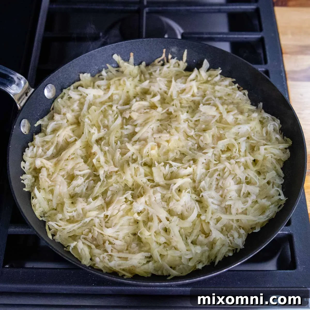 A close-up shot of potato rösti sizzling and browning in a non-stick skillet on the stovetop, showing the initial cooking stage.