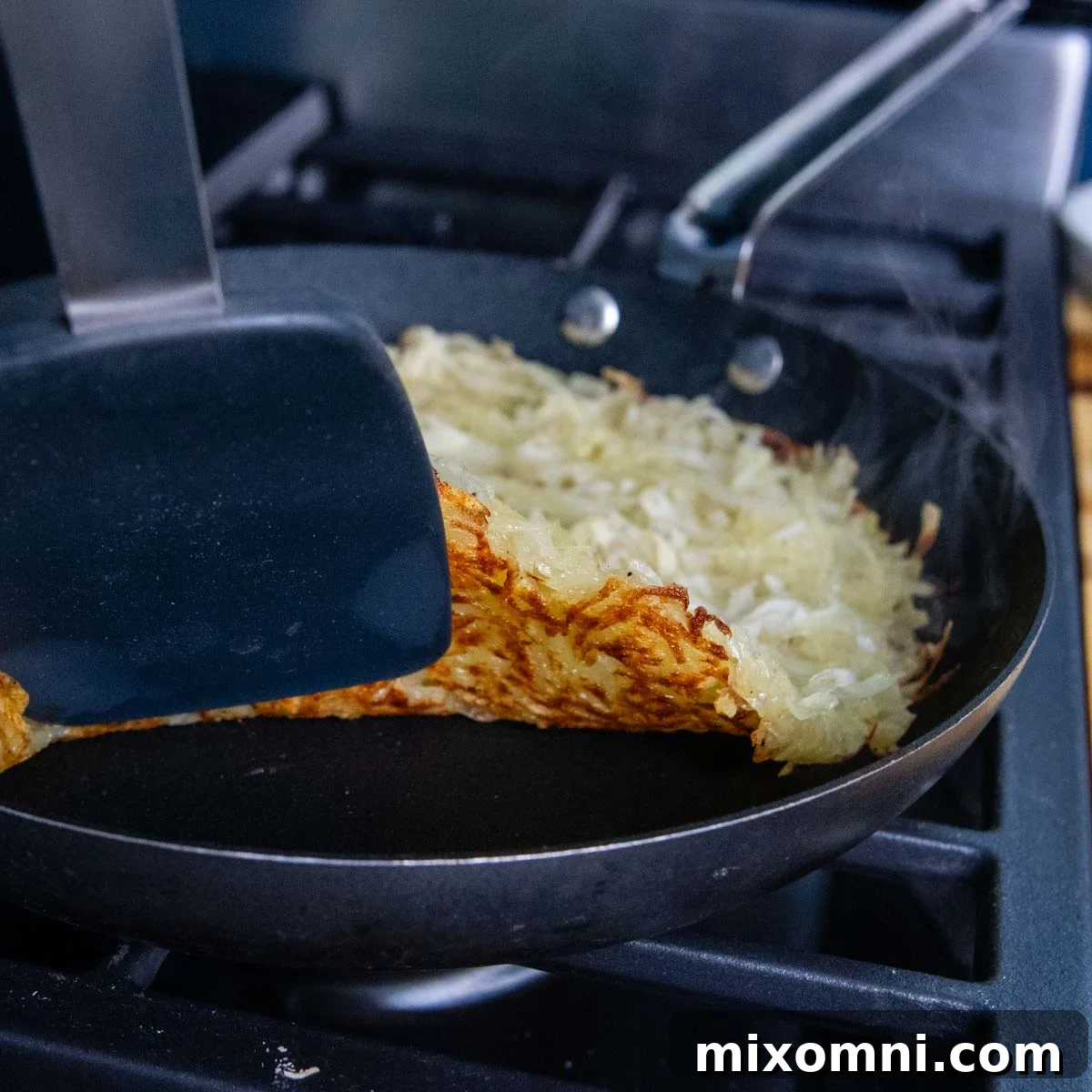 A spatula carefully lifting the edge of a potato rösti in a skillet, revealing a beautifully browned and crispy underside, indicating it's ready for flipping.