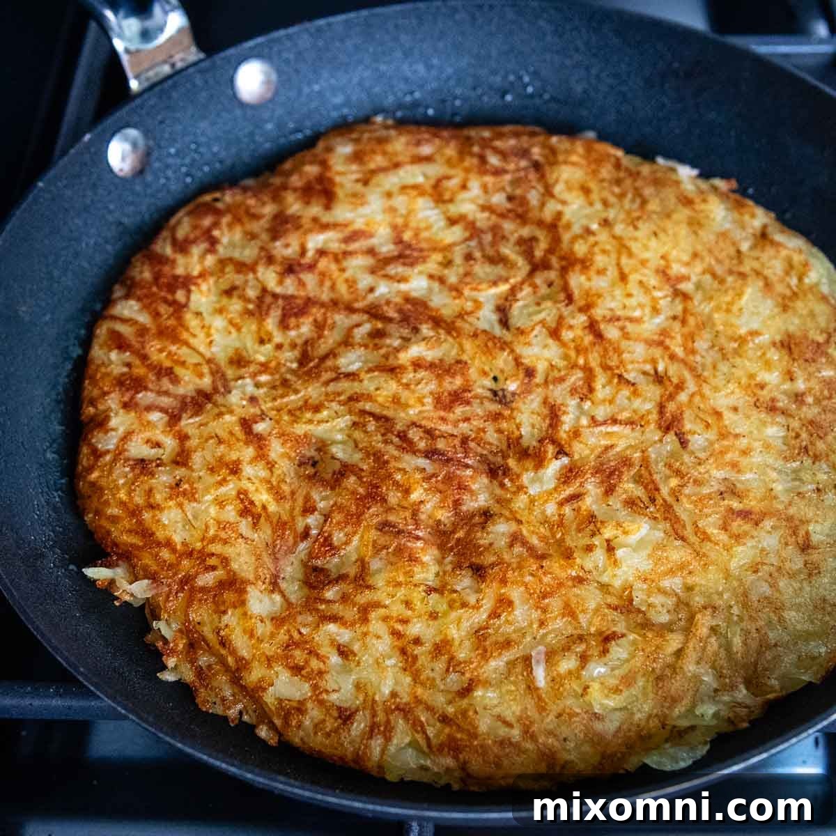 An overhead shot of a perfectly round, golden-brown potato rösti cooking in a non-stick skillet, showcasing its appealing texture.