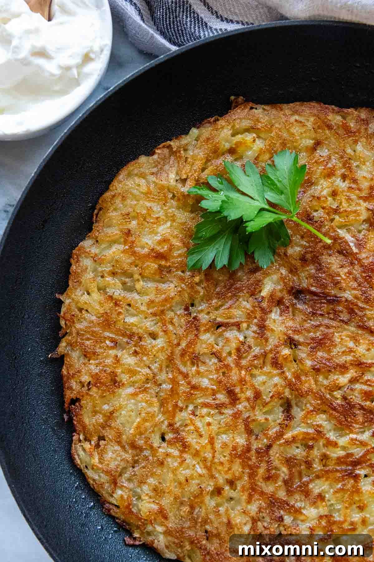 A golden-brown potato rösti resting in a black skillet, elegantly garnished with fresh chopped parsley, ready to be served.