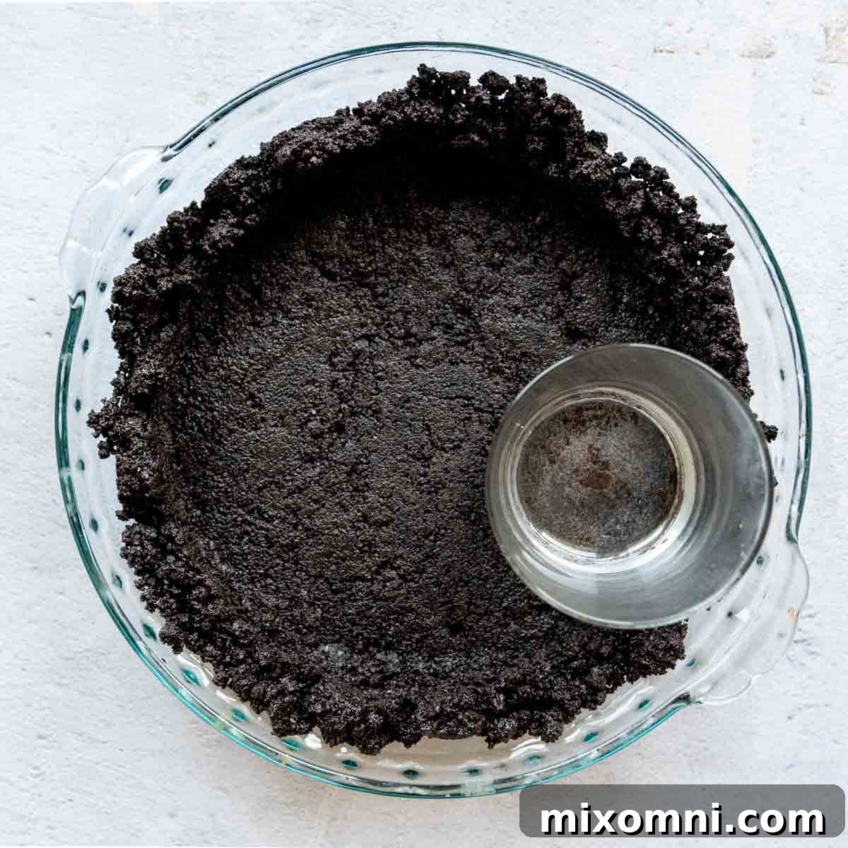 The gluten-free Oreo crust mixture being firmly pressed into the bottom of a glass pie dish using a drinking glass.