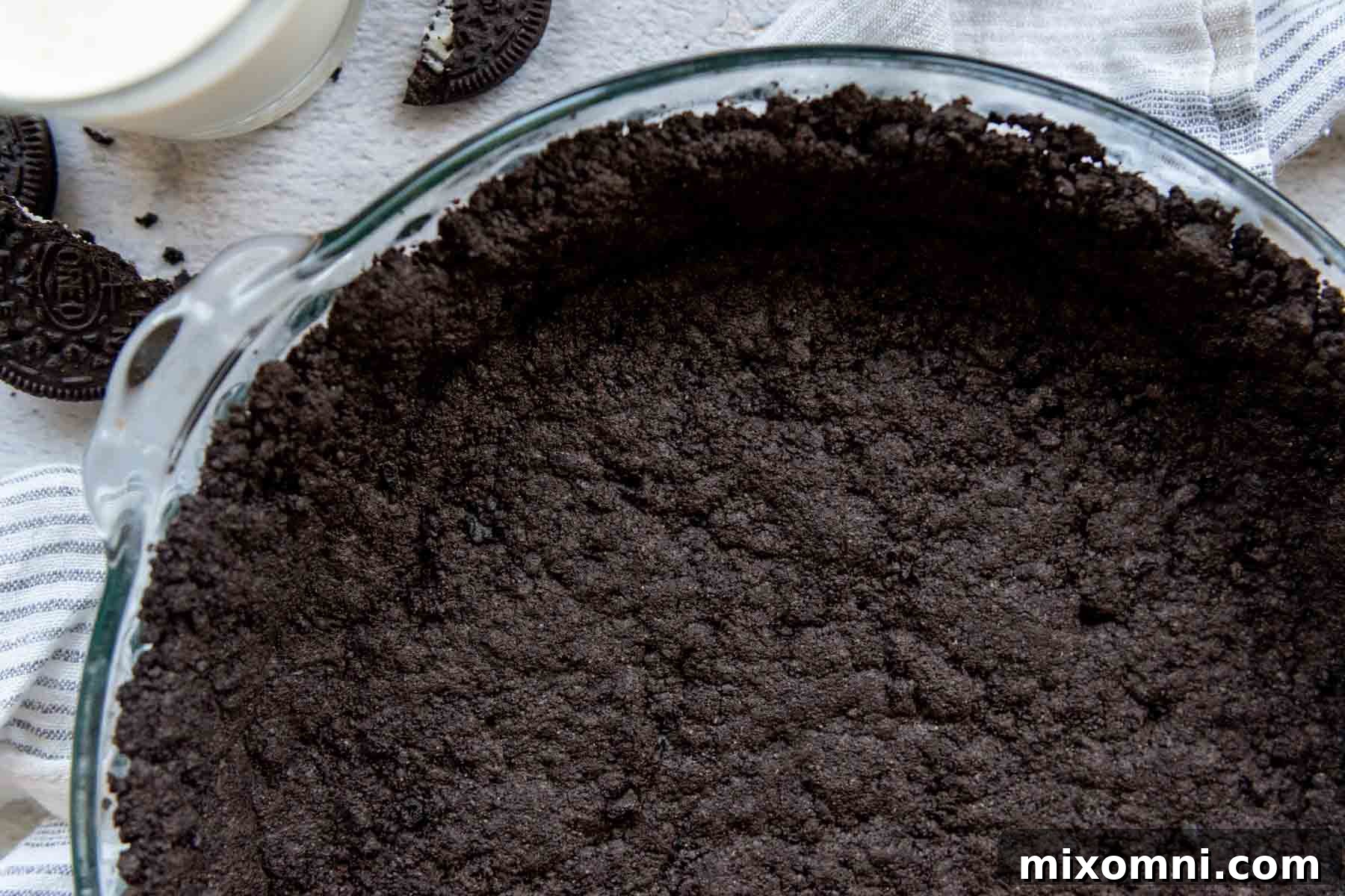 An overhead close-up shot of the beautifully baked gluten-free Oreo pie crust in a glass pie dish, with a glass of milk and two Oreos nearby, ready for indulgence.
