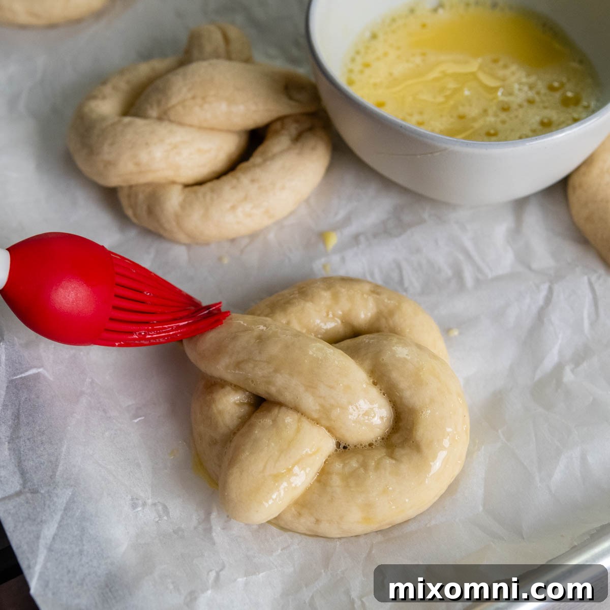 boiled pretzel laying on a piece of parchment paper being brushed with egg wash.