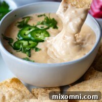 A tortilla chip being dipped into a bowl of gluten-free queso cheese sauce garnished with jalapeño.