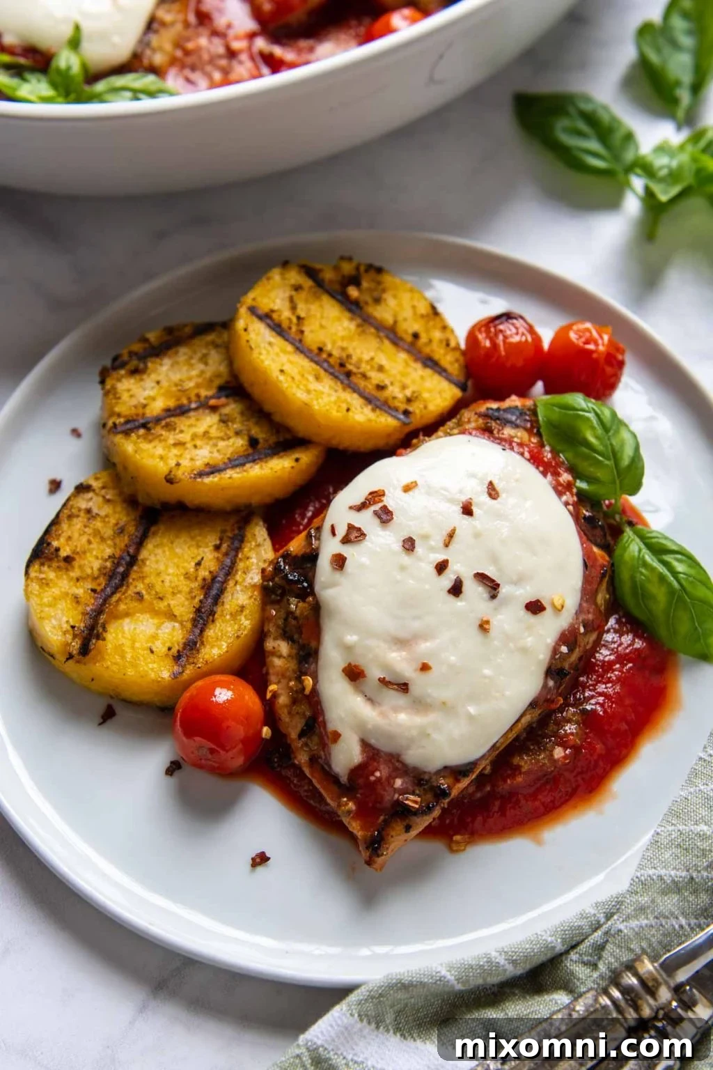 Close up of a plated grilled chicken parmesan dinner with fresh basil, with a large serving dish in the background