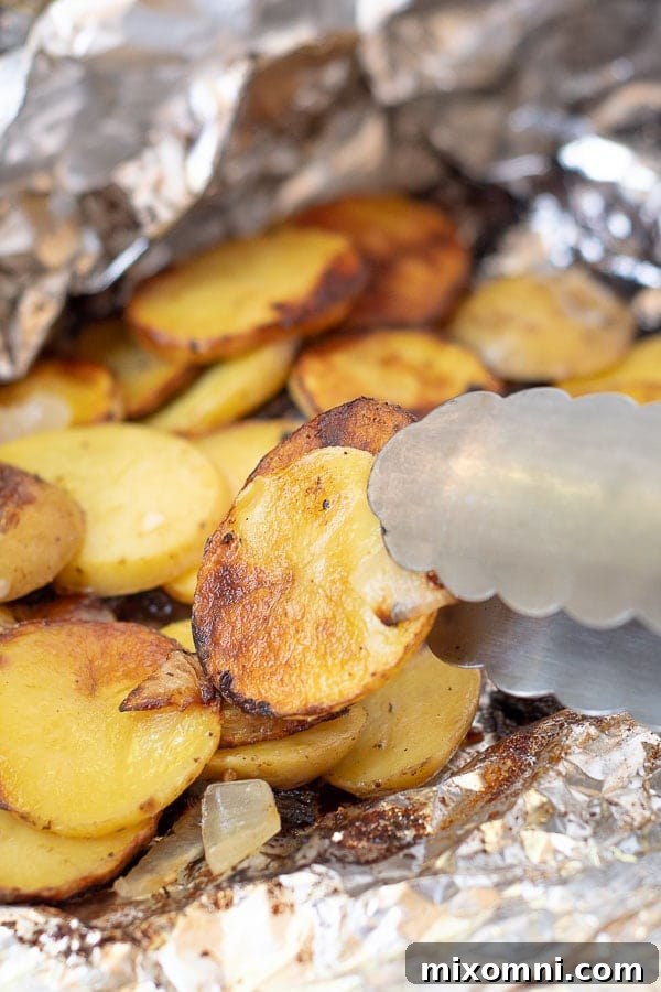 A pair of tongs carefully picking up a perfectly grilled potato slice from an open foil packet, showcasing its tender texture and golden-brown edges.