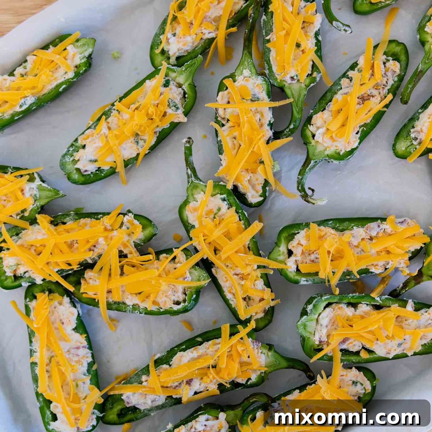 Jalapeño poppers on a baking sheet, filled with cream cheese and topped with extra cheese and bacon, ready for the oven.