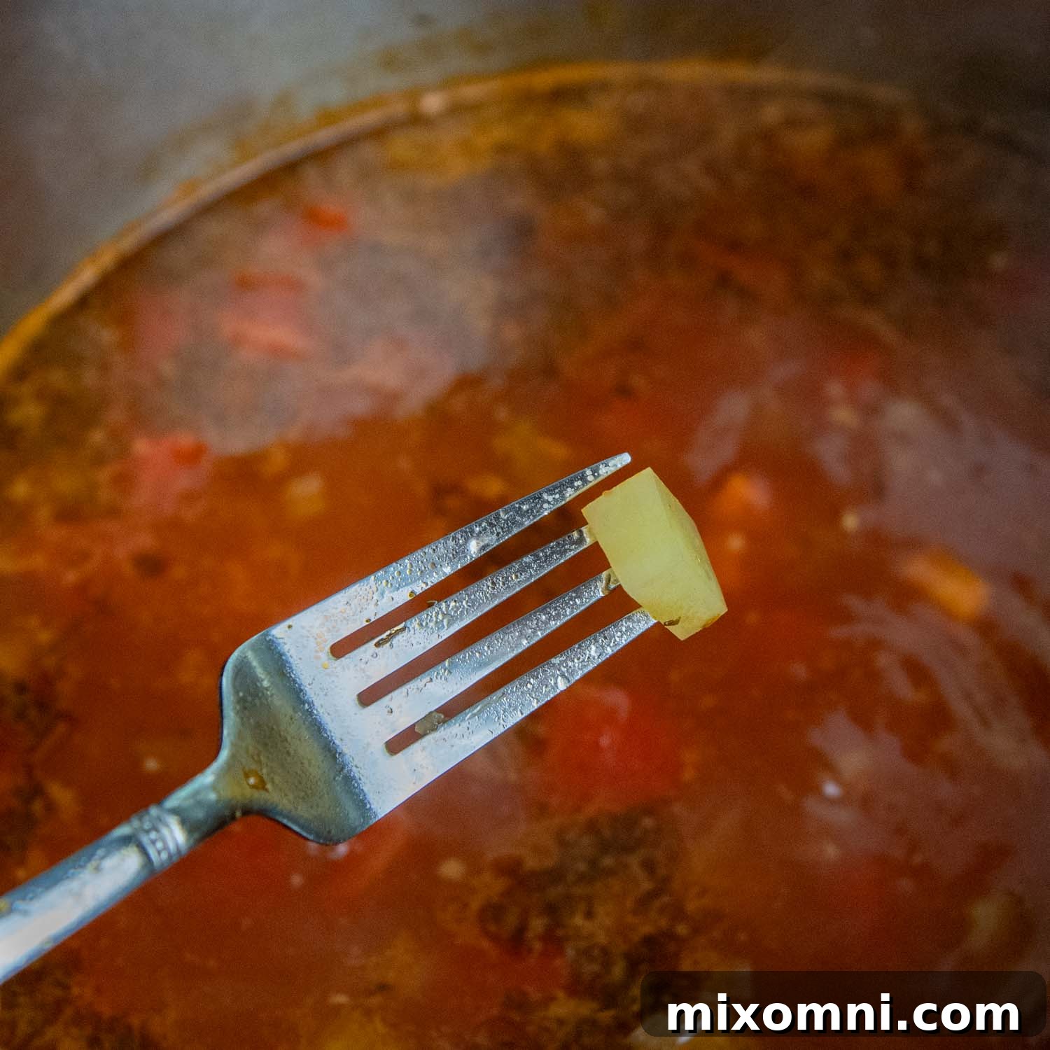 Fork testing the tenderness of a potato in a finished vegetable soup.