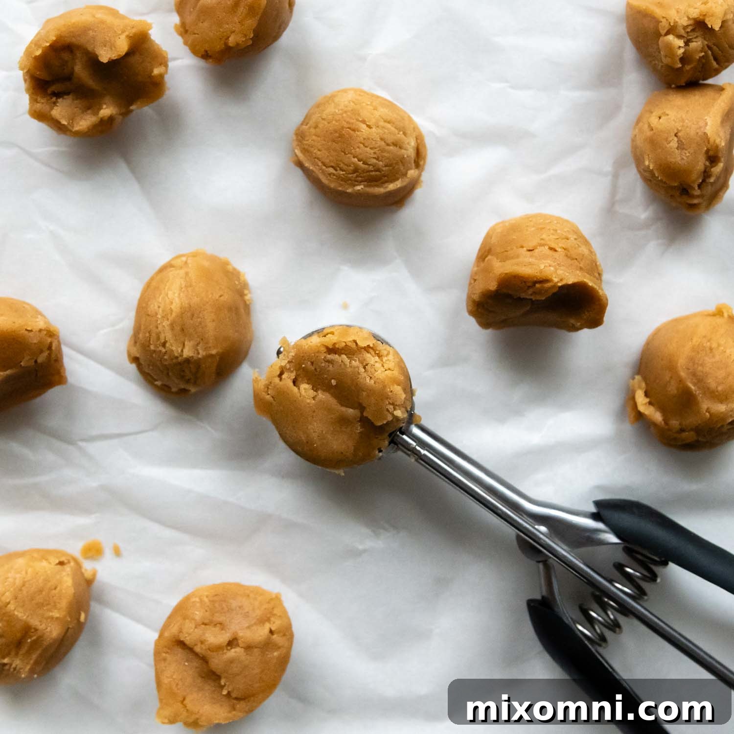 A small cookie scoop rests beside perfectly round cookie dough balls on parchment paper, ready for baking.