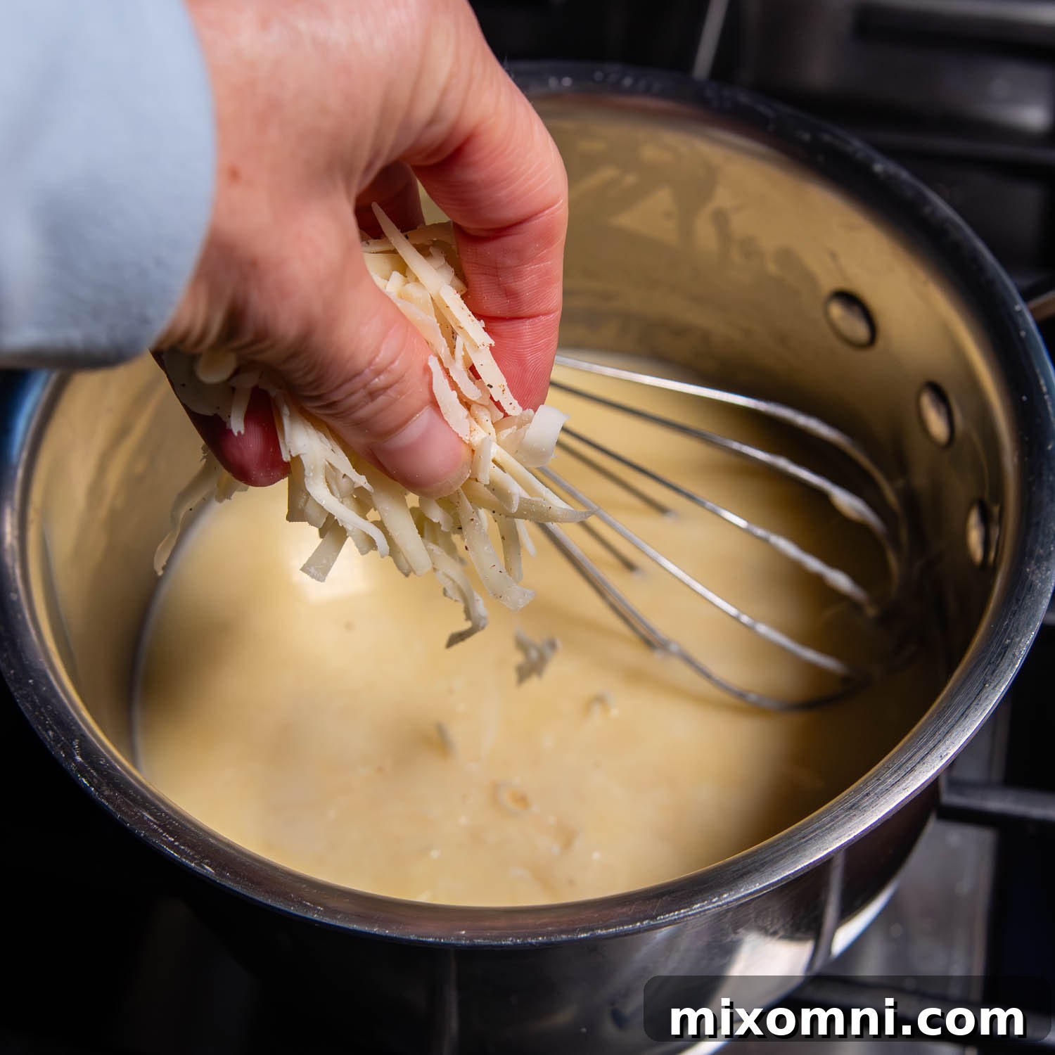 Hand adding shredded cheese to a pot with a whisk in it.