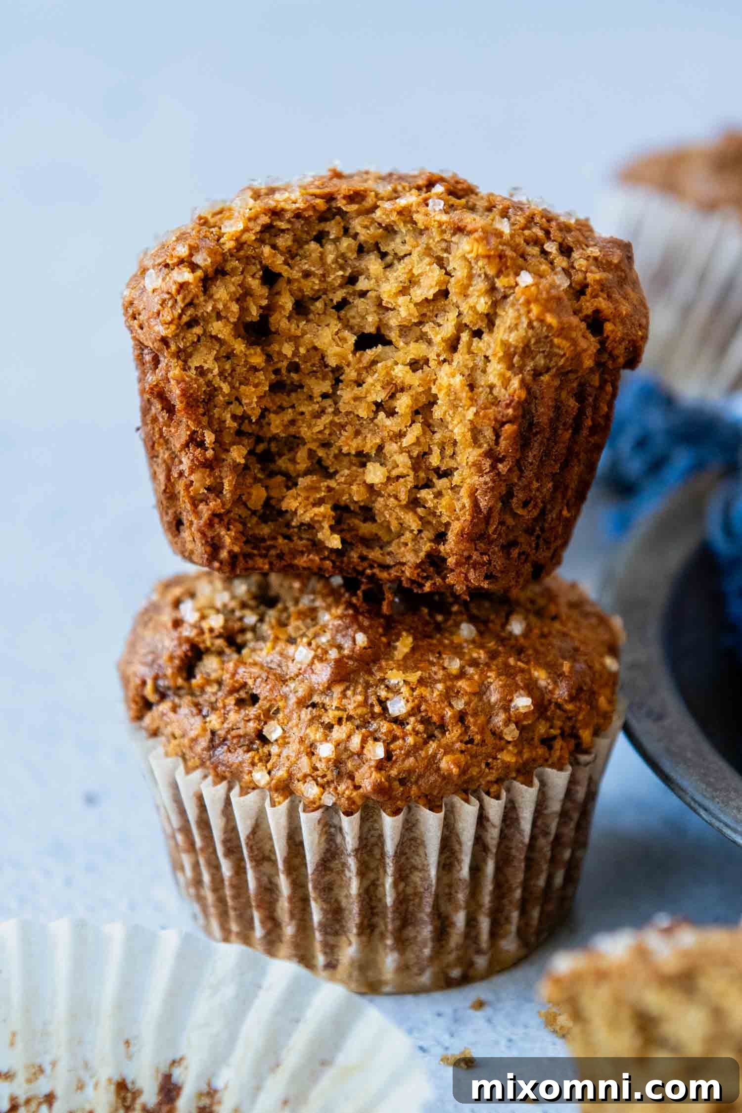 Two Gluten-Free Bran Muffins stacked with the top muffin having a bit taken out of it showing the inside texture. Rich, golden-brown tops with a sprinkle of coarse sugar, showcasing a tender, moist interior dotted with wholesome oat bran.
