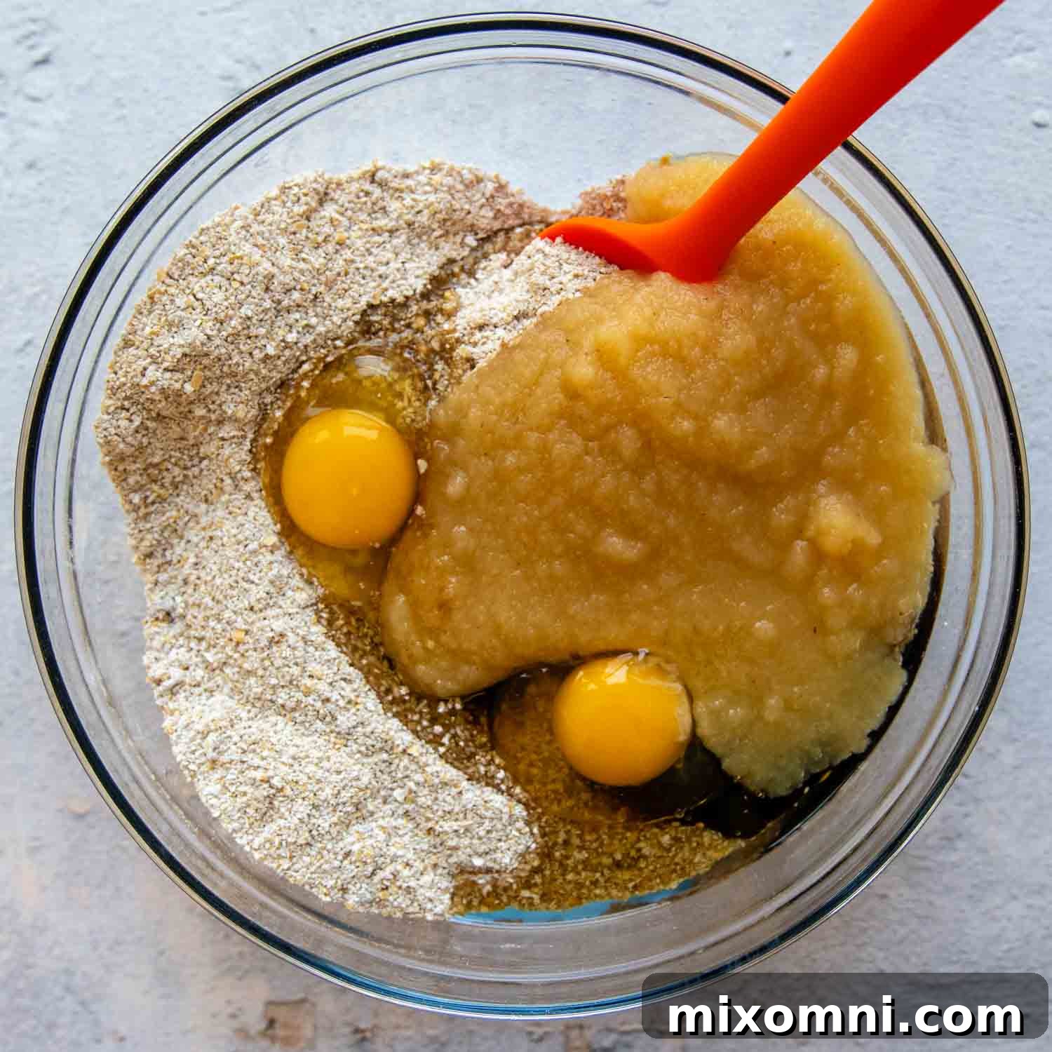 Wet and dry muffin ingredients in a large glass bowl with a red spatula. The batter is thick and well-combined, ready for baking.