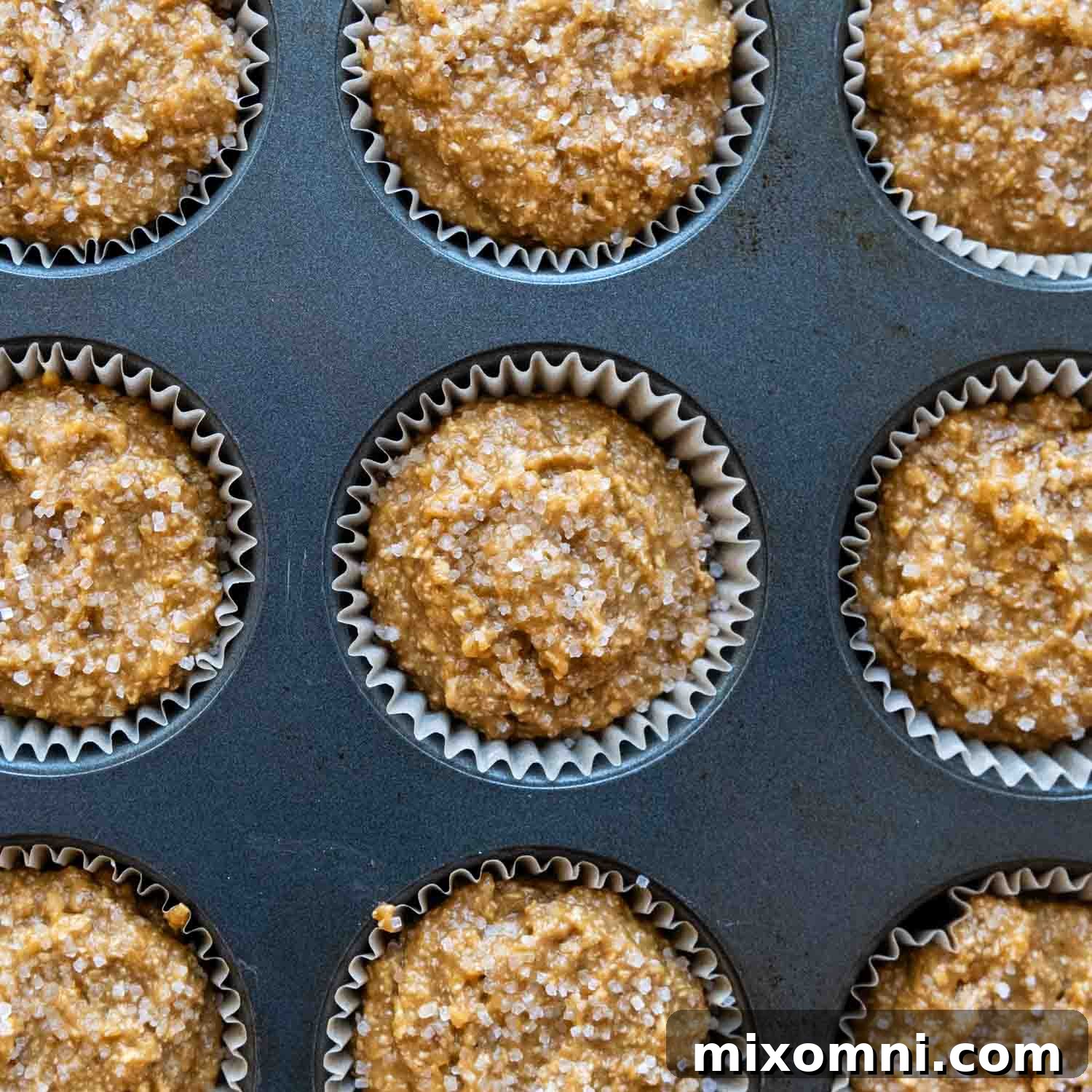 Uncooked muffin batter in muffin tin, sprinkled with coarse sugar, resting before baking.