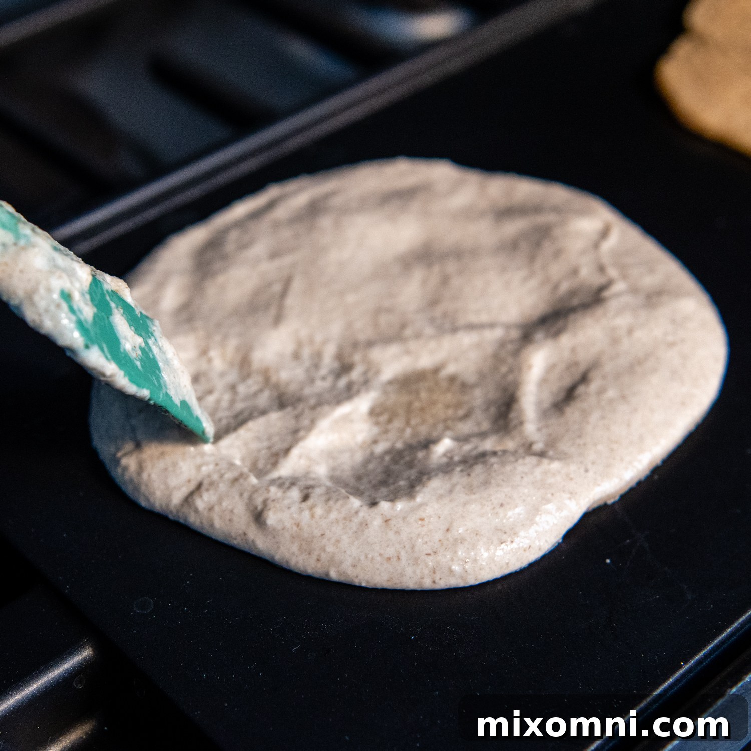 A silicone spoon expertly spreading thin oat tortilla batter in a circular motion on a hot griddle.