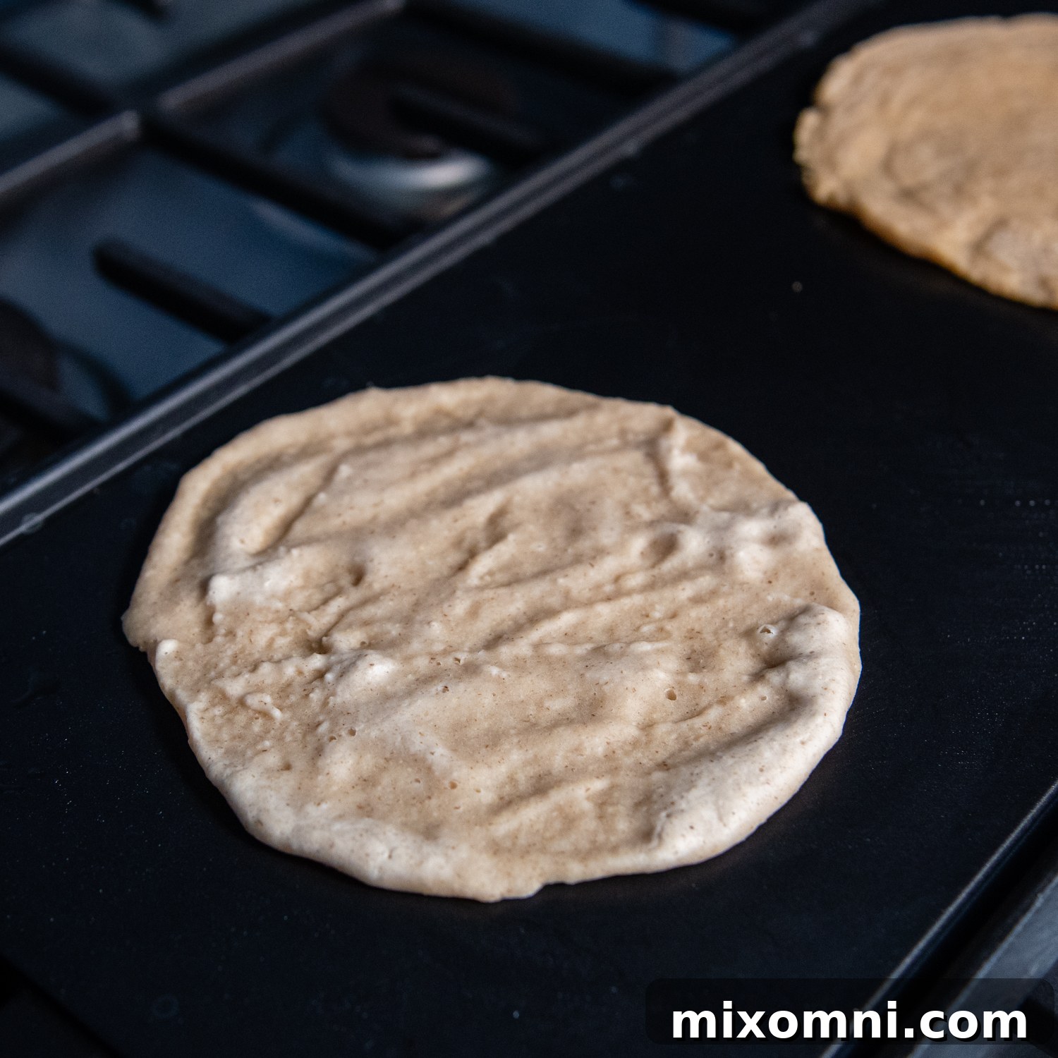 A perfectly circular, thin oat tortilla spread evenly on the griddle, just beginning to cook.