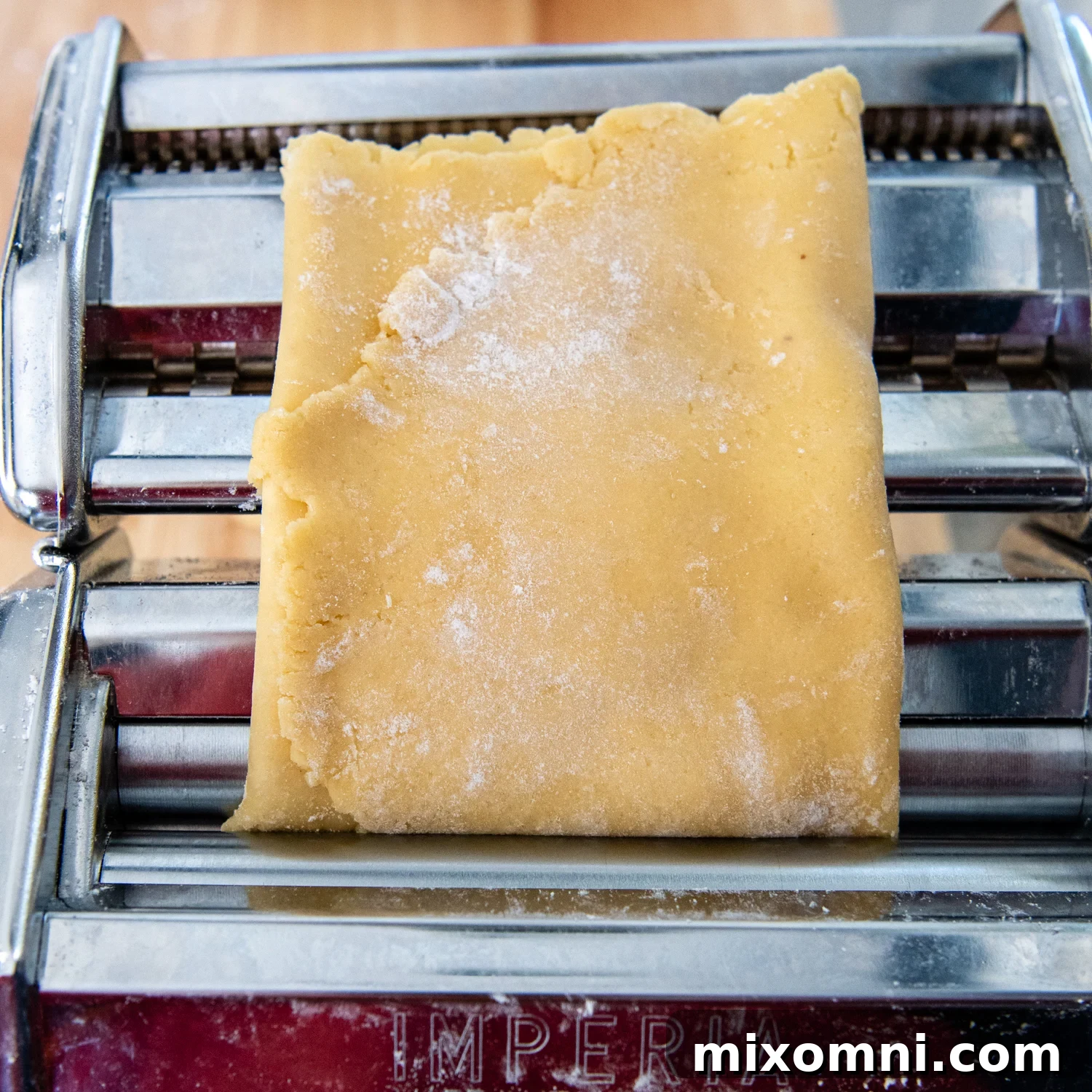 Gluten-free pasta dough, now smoother, being fed through the widest setting of the pasta machine after a second fold and turn.