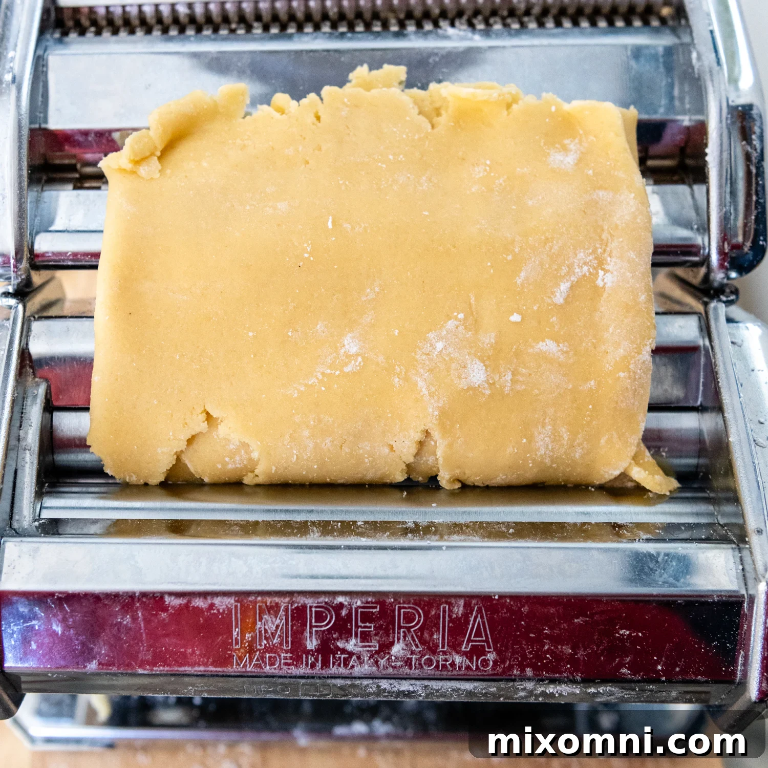 Folded gluten-free pasta dough being fed into the widest setting of a pasta machine for a second pass.