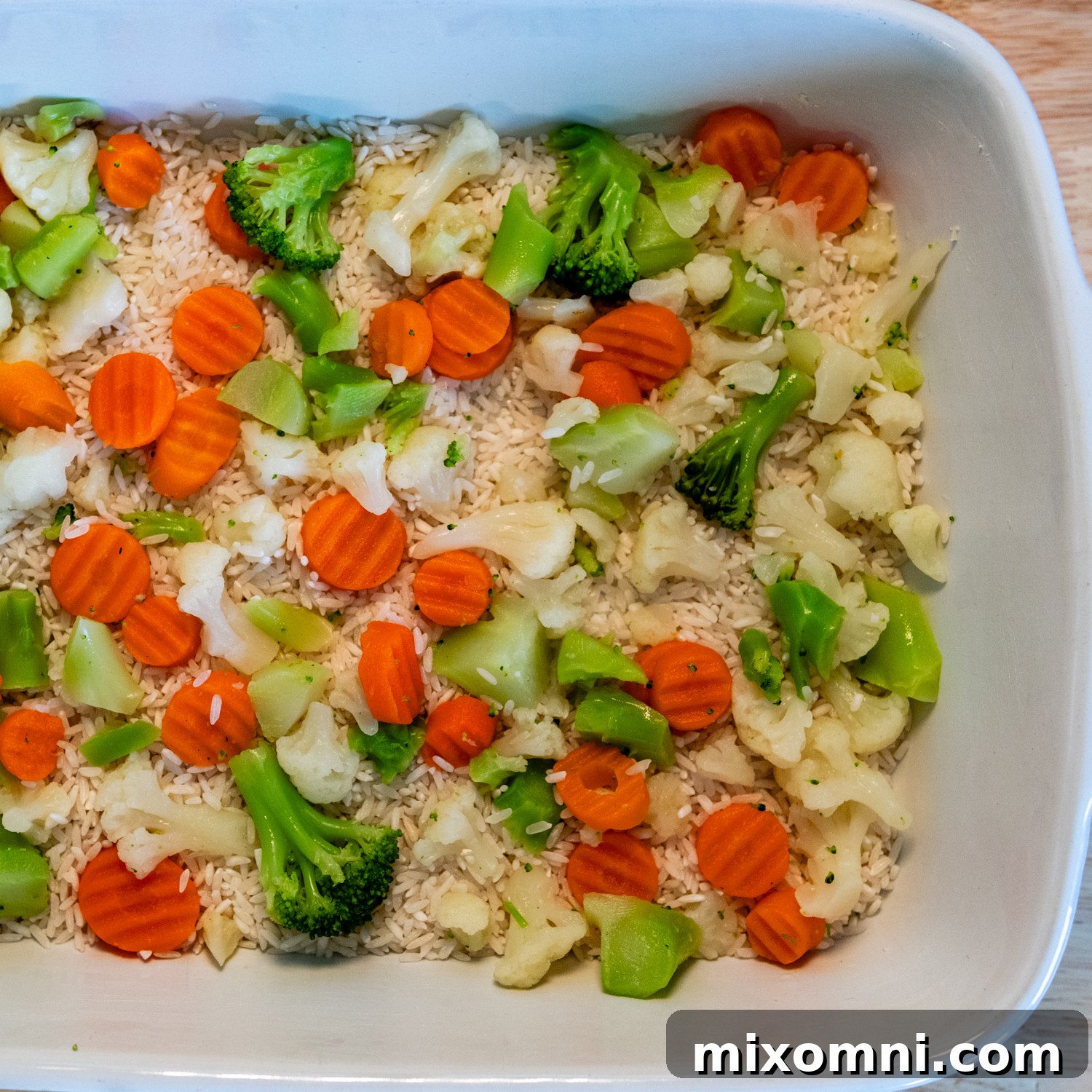 Layers of uncooked rice, frozen vegetables, and seasoned raw chicken breast in a white baking dish, ready for sauce.