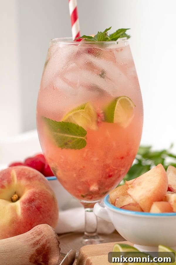 A Raspberry Peach Mojito in a tall glass with cut up peaches and muddler next to it