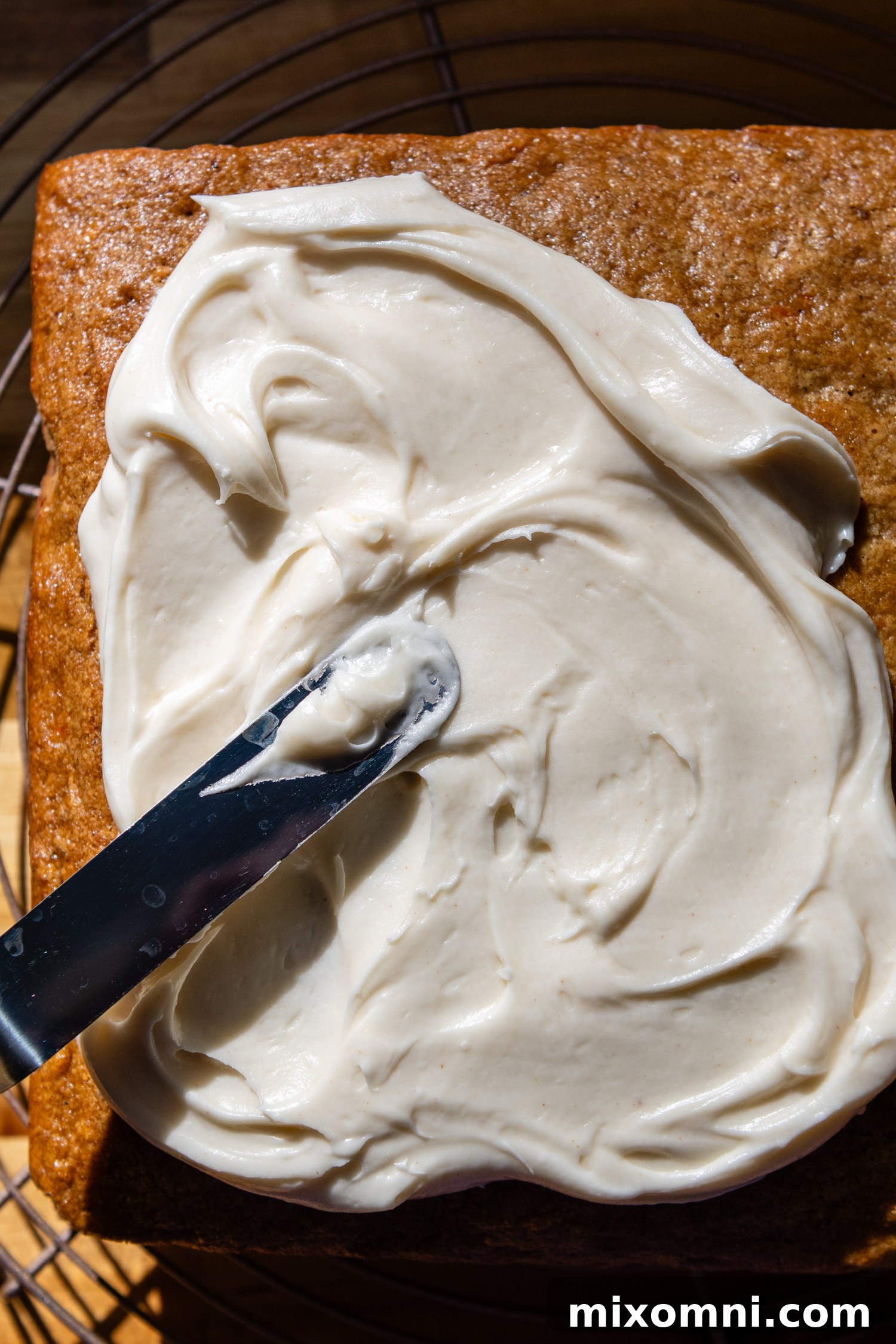A knife spreading a thick, creamy layer of browned butter cream cheese frosting smoothly over the cooled oat flour carrot cake.