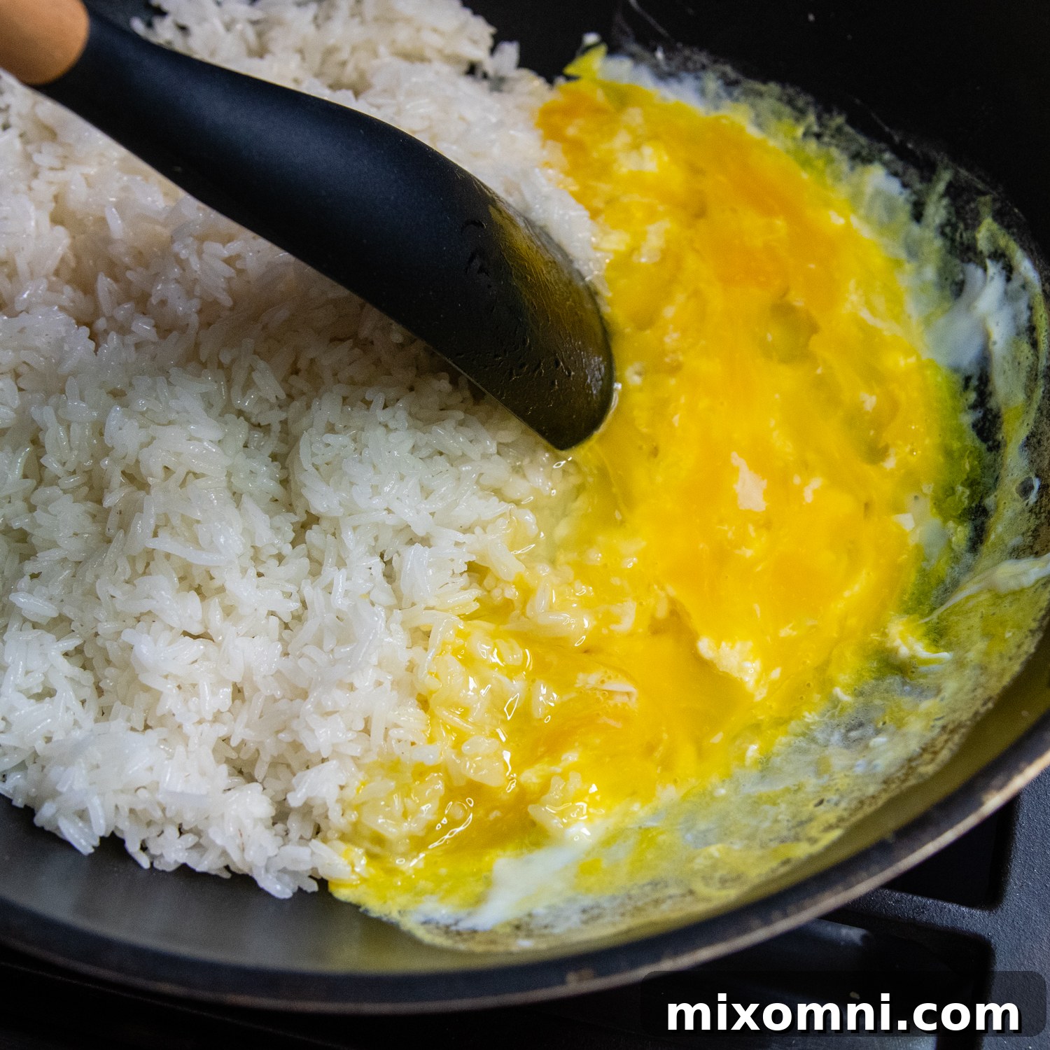 Scrambled eggs being mixed with toasted rice in a black wok using a spatula, showing the combination of ingredients.