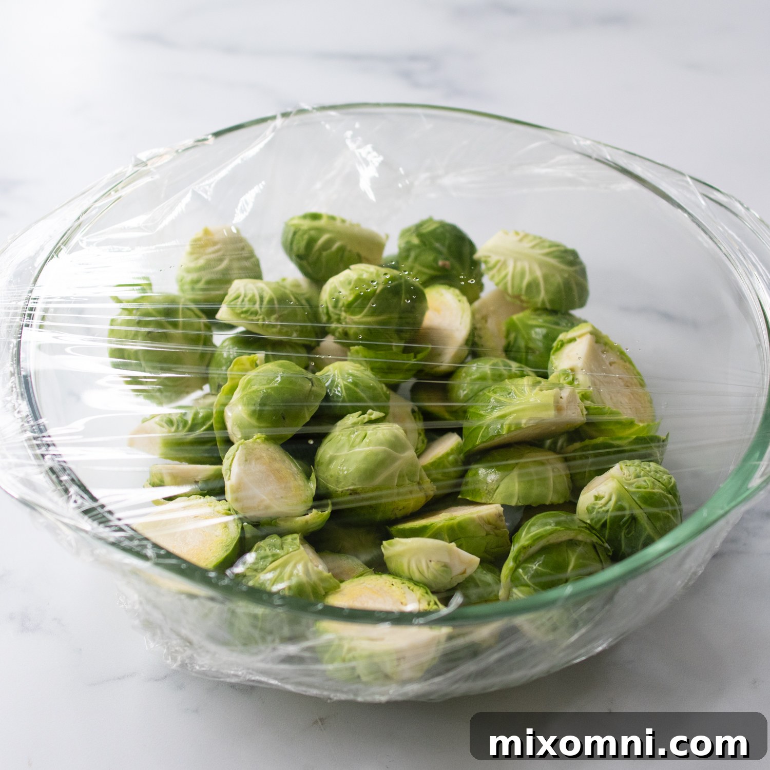 A bowl of Brussels sprouts in a glass bowl covered with cling wrap, ready for microwaving to par-steam them.
