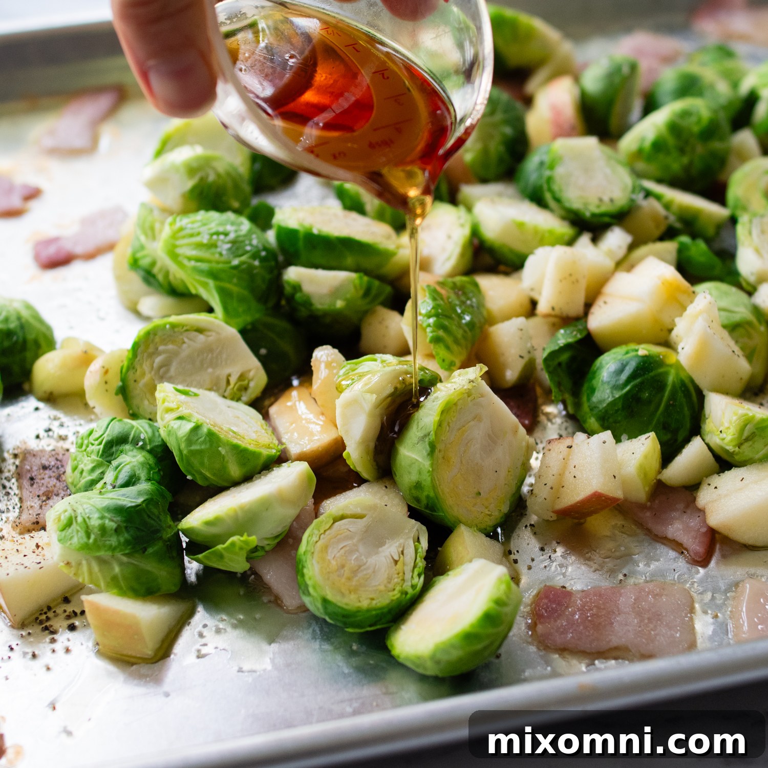 Brussels sprouts arranged on a sheet pan with maple syrup being drizzled over them, ready for roasting.