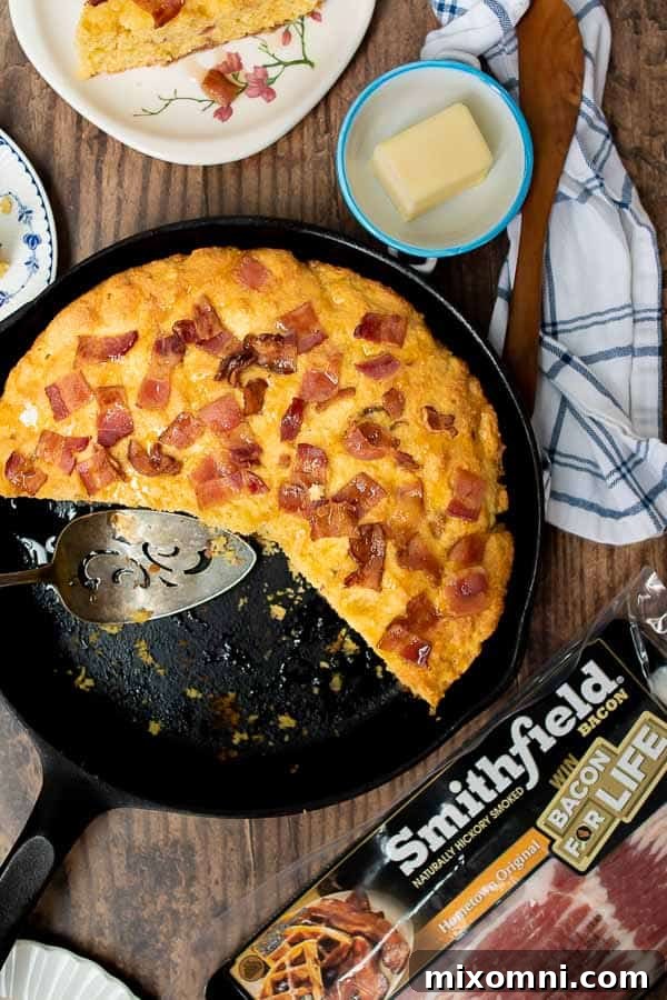 An overhead shot of a freshly baked bacon cornbread in an iron skillet, with a slice removed and small plates ready for serving around it. The golden crust hints at its delicious maple-bacon topping.