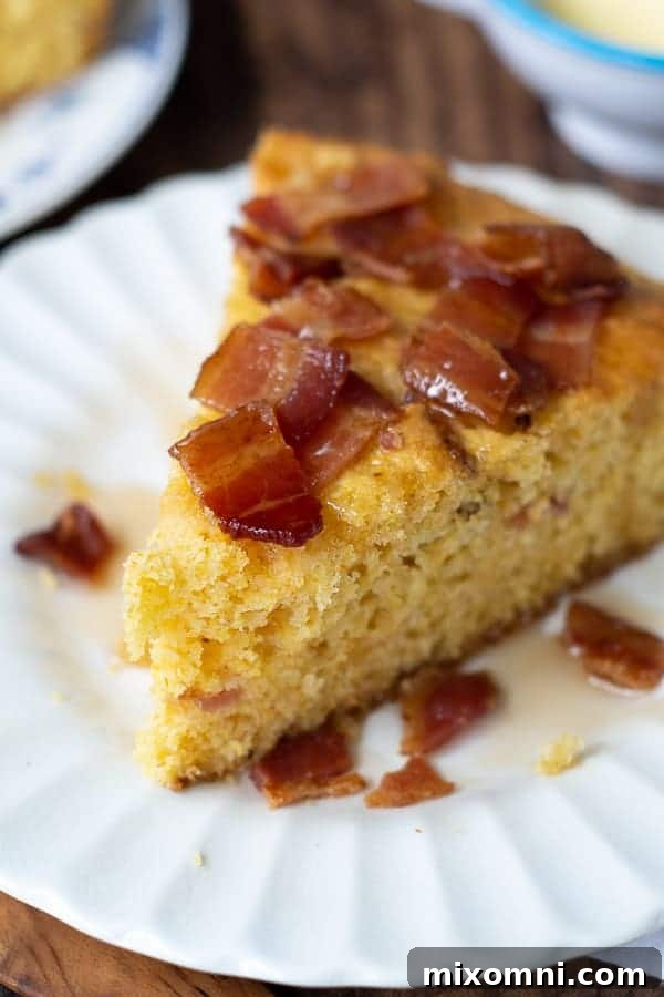 A perfectly cut wedge of bacon maple cornbread rests on a white plate, glistening with a drizzle of syrup. The golden-brown crust and visible bacon pieces promise a delightful sweet and savory experience.