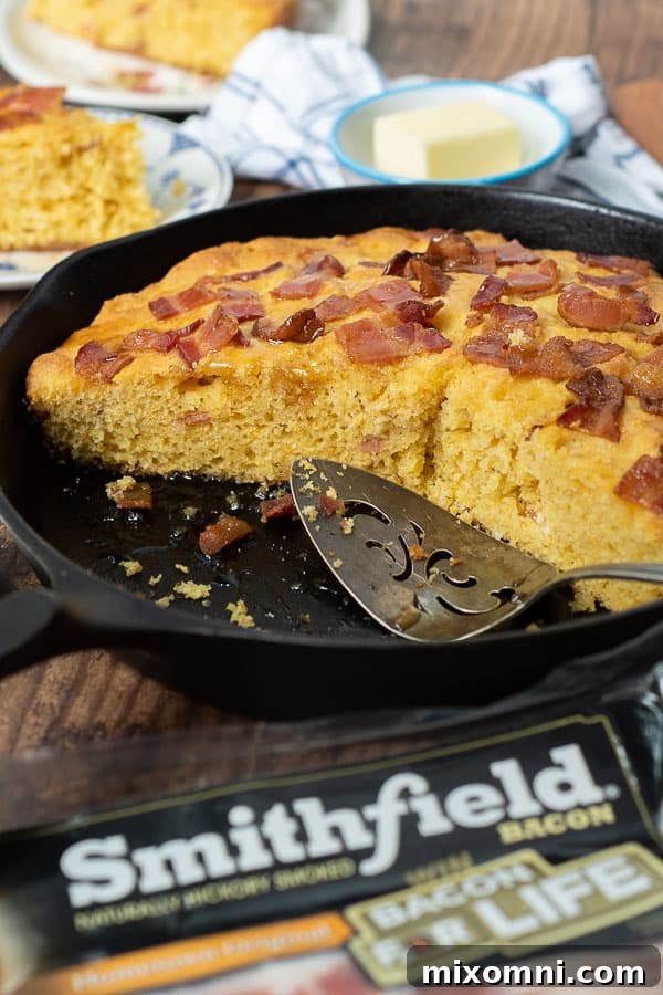 A cast iron skillet filled with delicious bacon cornbread, featuring several slices already cut and removed, showcasing the golden-brown crust and moist interior.