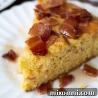 A wedge of bacon maple cornbread on a white plate with syrup drizzled over, a perfect blend of sweet and savory.