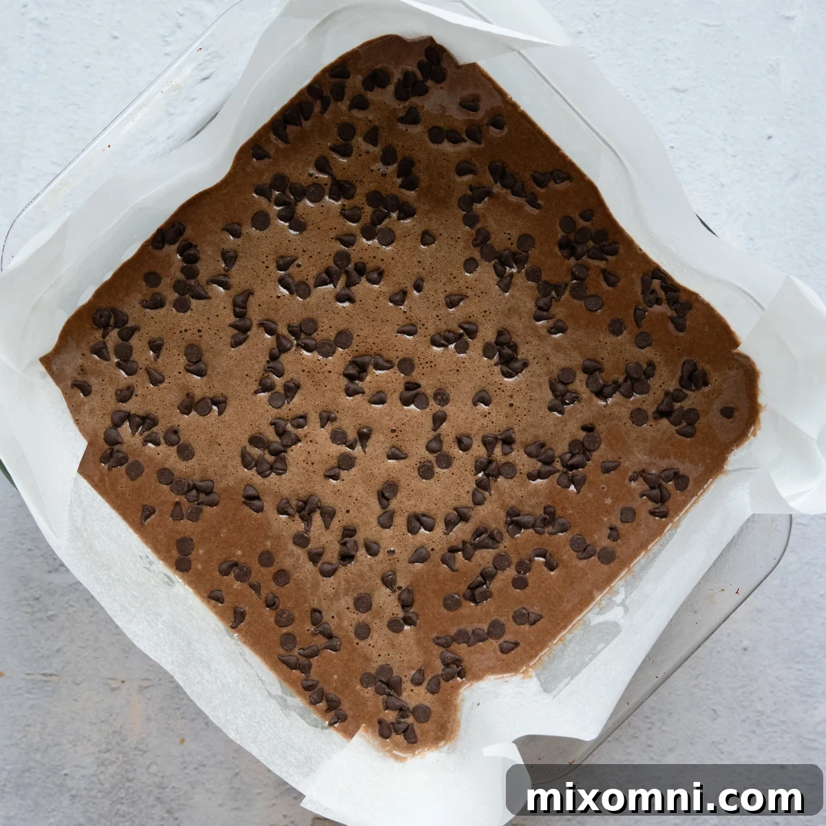 Chocolate cake batter for oat flour chocolate cake poured into a greased 8-inch baking dish, topped with extra chocolate chips.