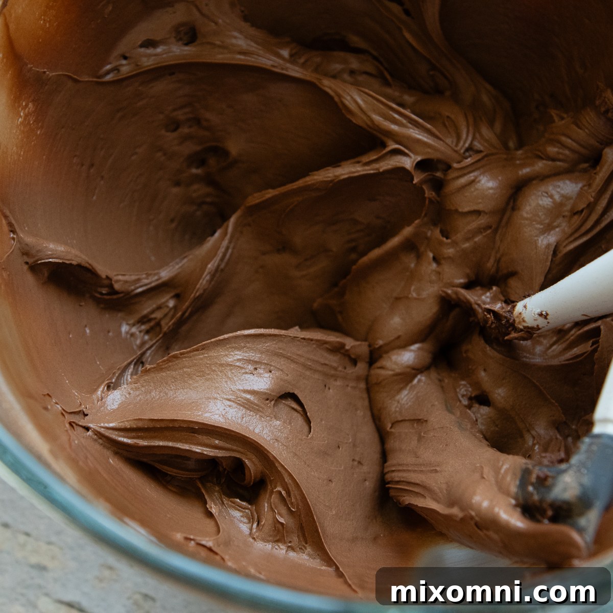 Rich, homemade chocolate frosting in a clear glass bowl, being stirred with a white spatula.