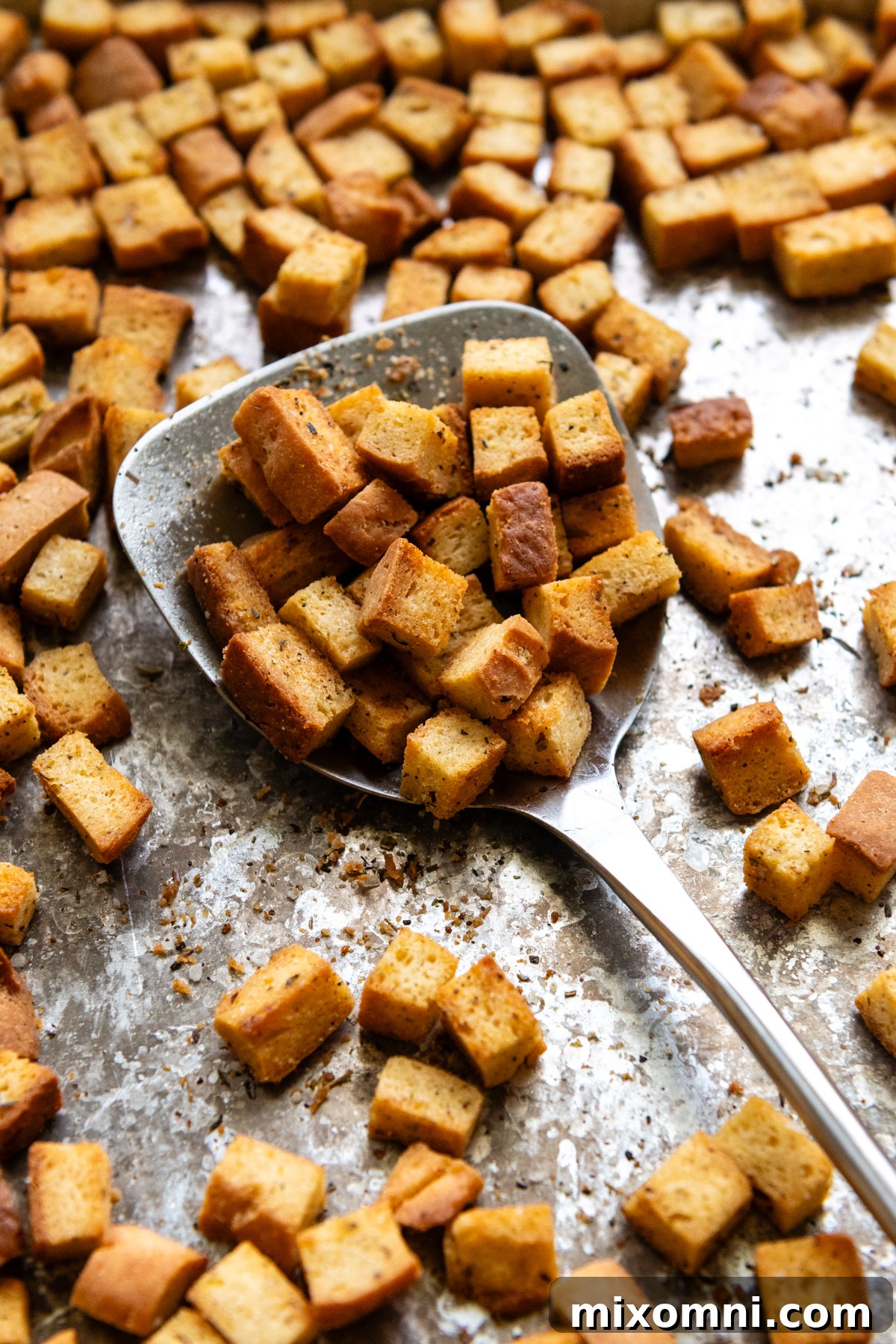 A metal spoon rests on a baking pan filled with golden-brown, crispy gluten-free croutons, fresh from the oven.