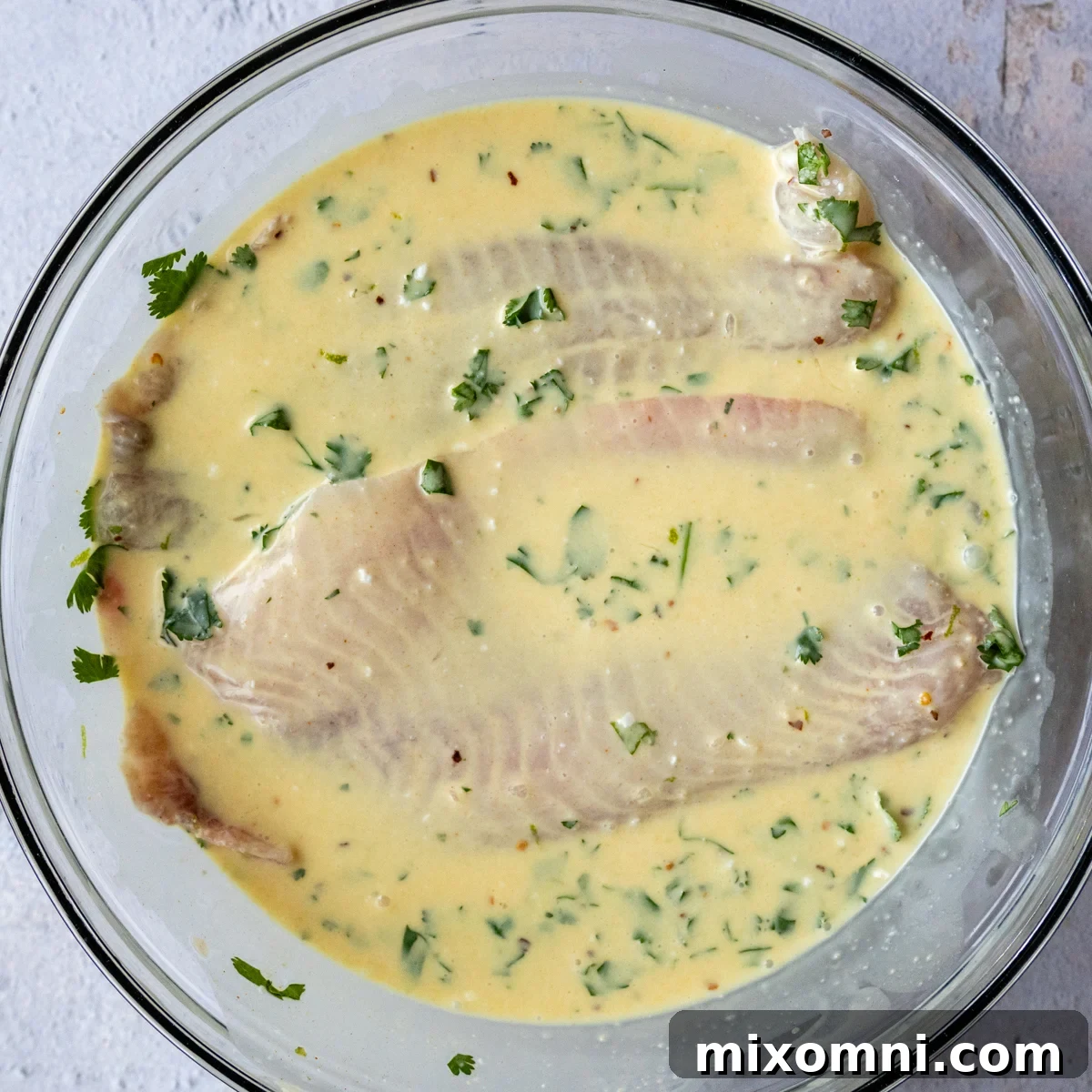 White fish fillets marinating in a creamy coconut milk sauce in a clear glass bowl, ready for baking.