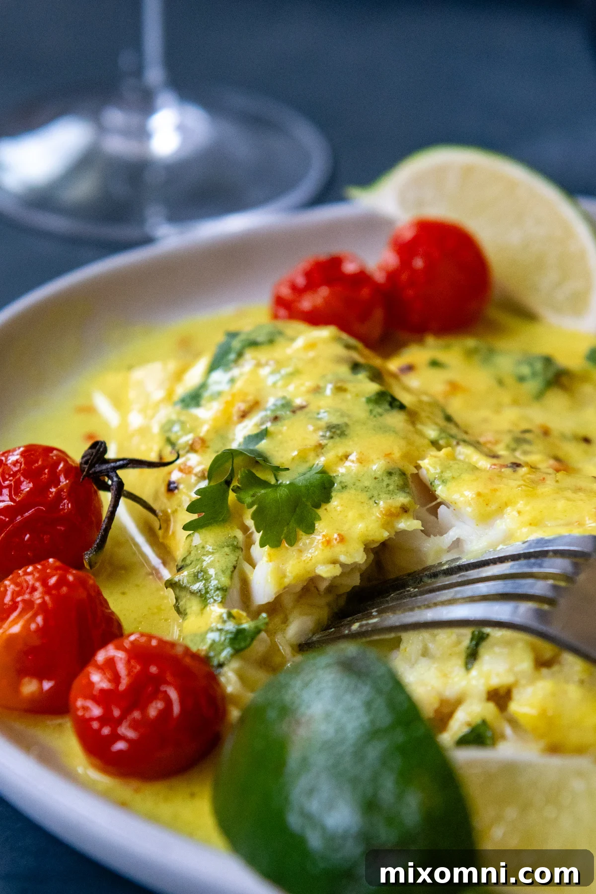 Cooked white fish flaking easily with a fork on a white plate, served alongside blistered cherry tomatoes and a rich coconut sauce.