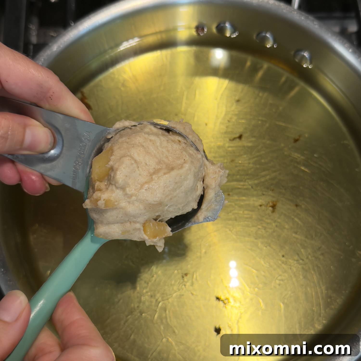 A measuring cup holding a doughy fritter over pot of oil.