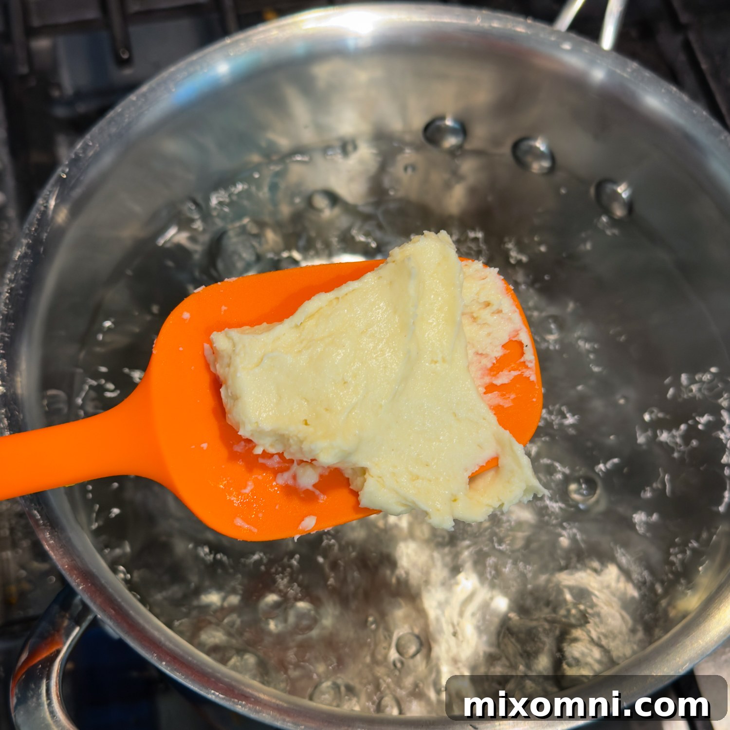 A vibrant orange spatula holding a dollop of dumpling dough over boiling water, ready to be dropped.