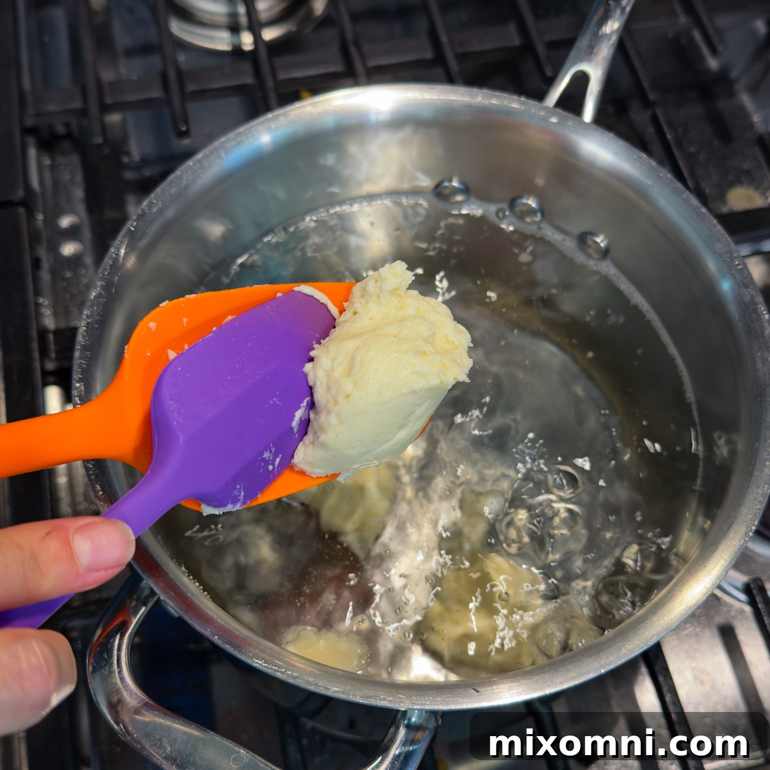 A purple spatula gently pushing dumpling dough from an orange spatula into bubbling boiling water.