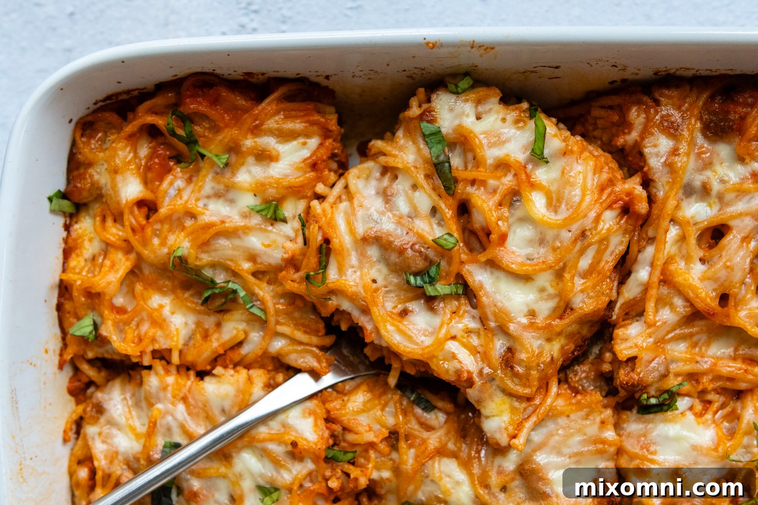 A serving of golden, cheesy gluten-free spaghetti casserole held by a spatula within the white baking dish, ready to be enjoyed.
