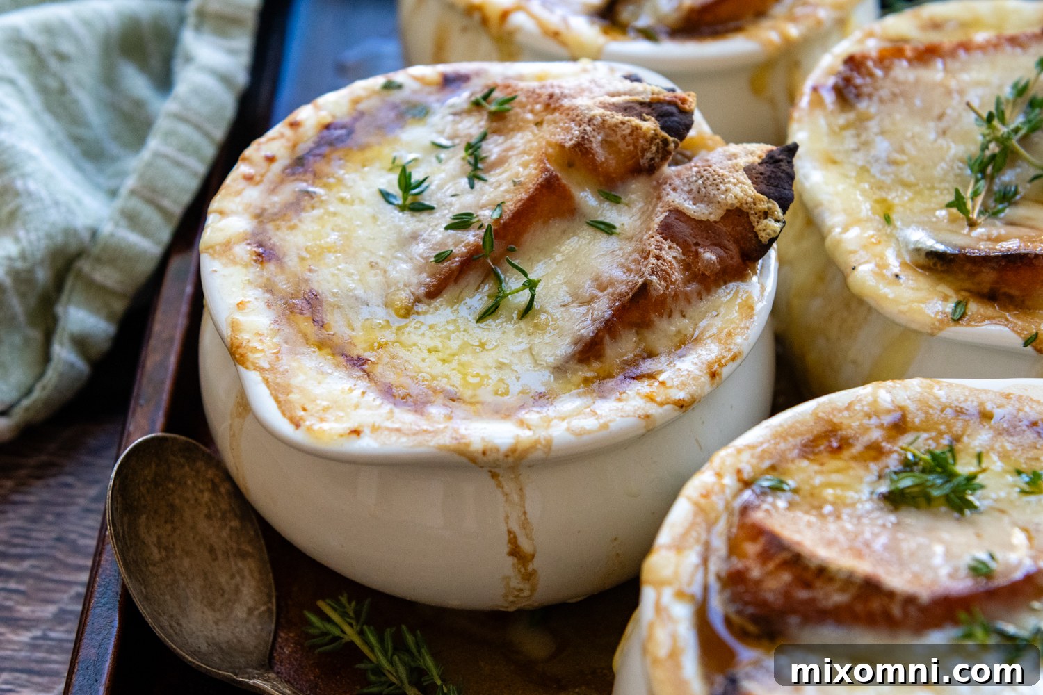 A close-up shot of a white soup crock filled with Gluten-Free French Onion Soup, topped with a toasted gluten-free French bread slice and melted, golden-brown Gruyere cheese, garnished with fresh herbs.