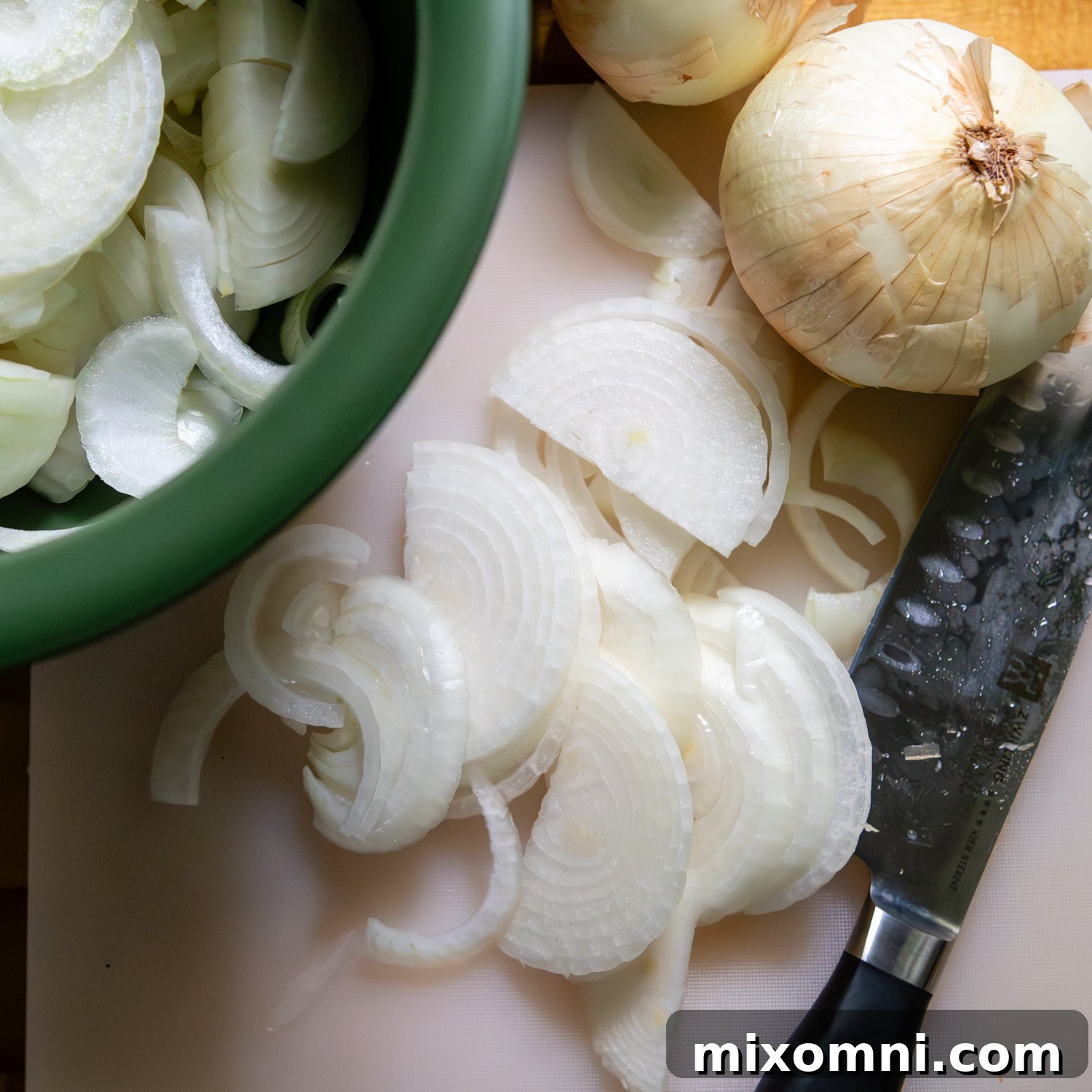 Green bowl filled with thinly sliced Vidalia onions, ready for cooking, with whole onions beside it on a cutting board.