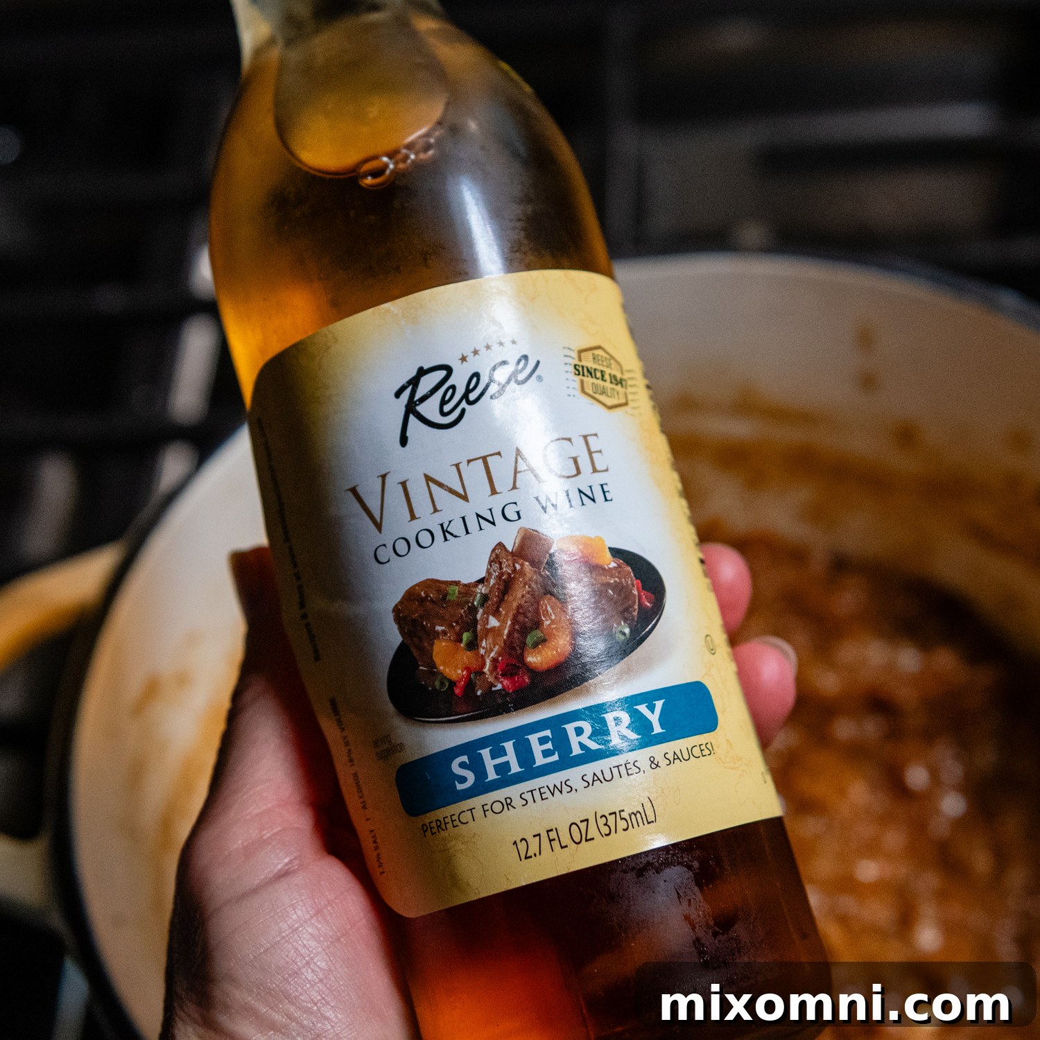 A hand holding a bottle of dry cooking sherry, ready to be added to the caramelized onions.