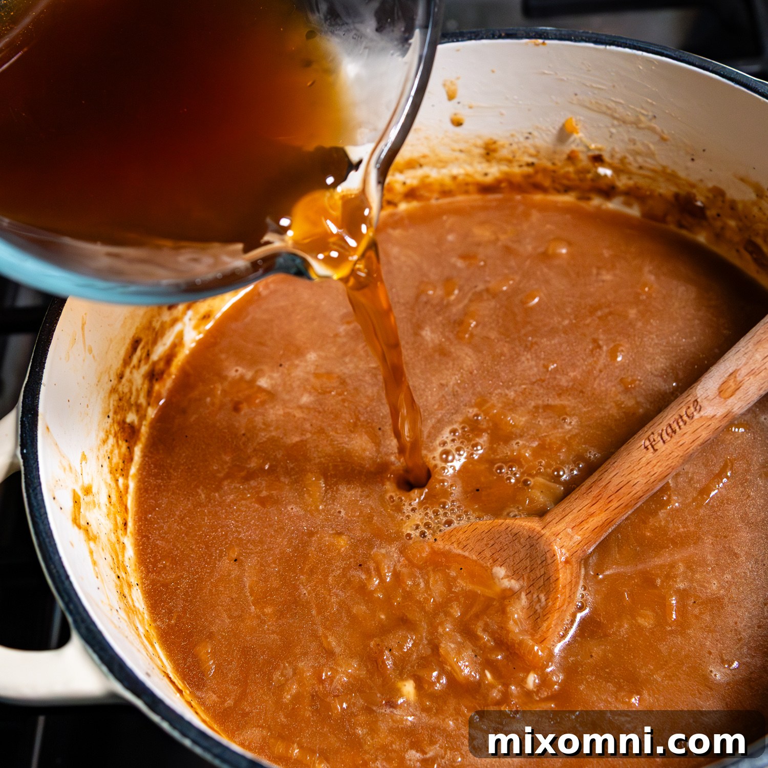 Gluten-free beef broth being poured into a pot of cooked, deeply caramelized onions, ready to form the soup's base.