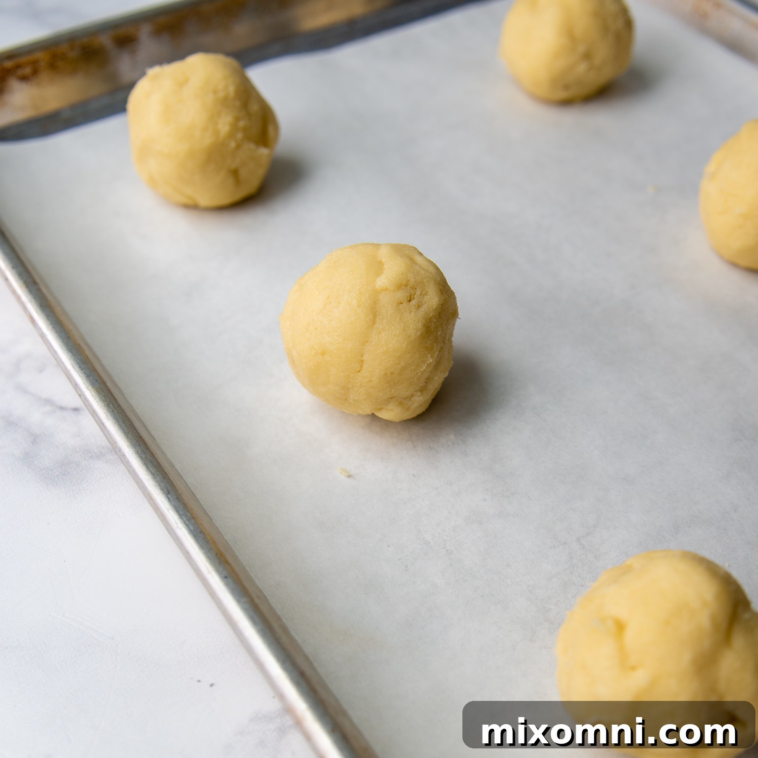 Formed cookie dough balls, flattened with a well in the center, on parchment paper on a baking sheet, ready for the oven.