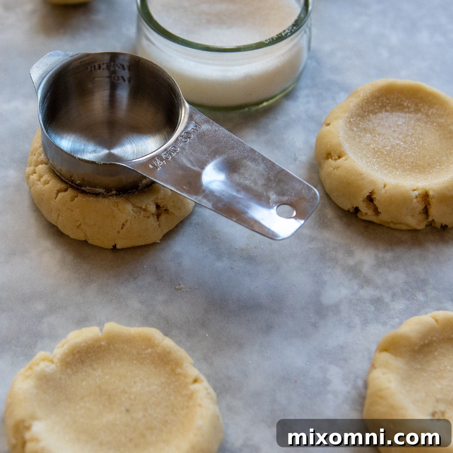 A measuring cup, dipped in sugar, being pressed into the center of a cookie dough ball to create an indentation.