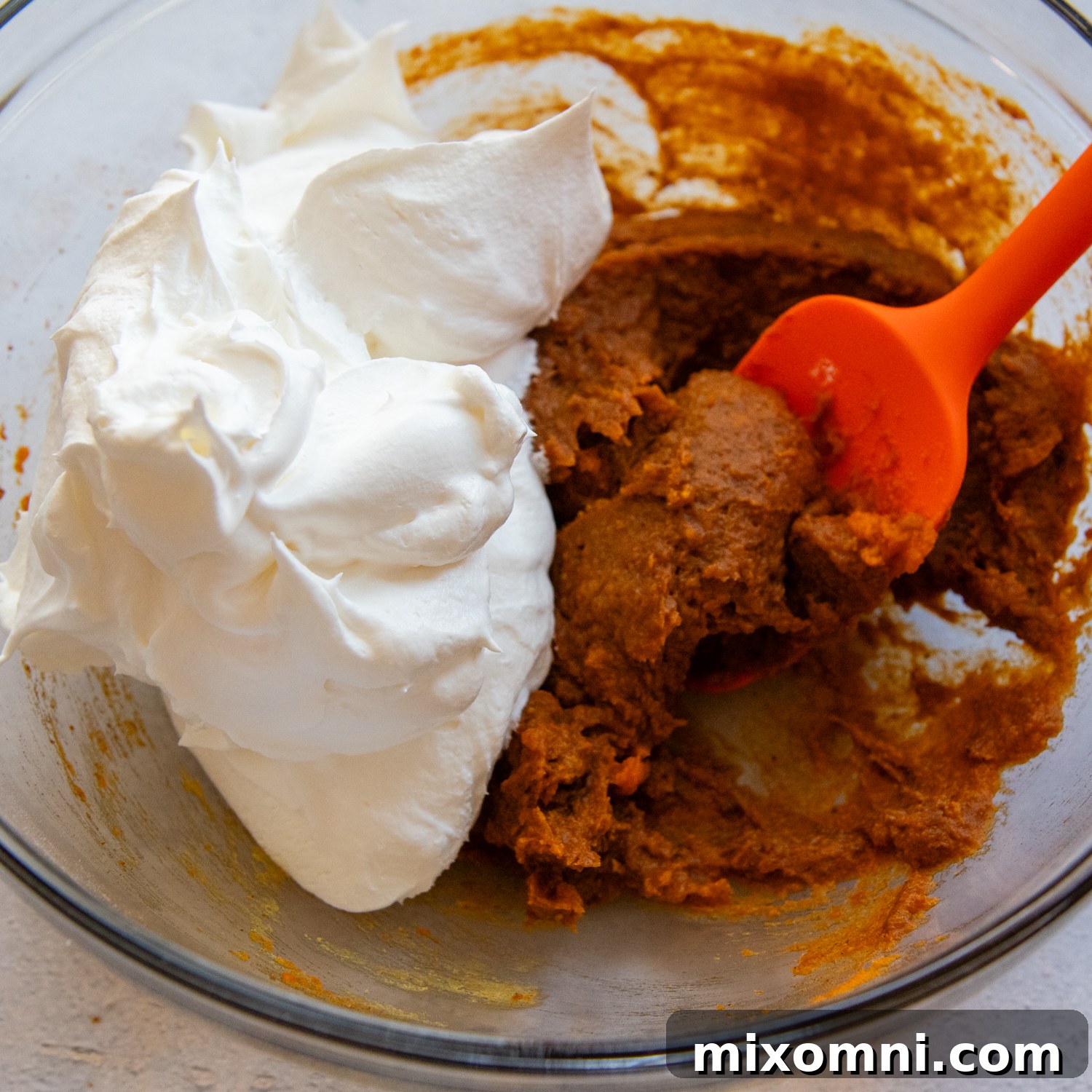 Canned pumpkin, instant pudding mix, and pumpkin pie spice being combined in a glass mixing bowl with a red spatula.