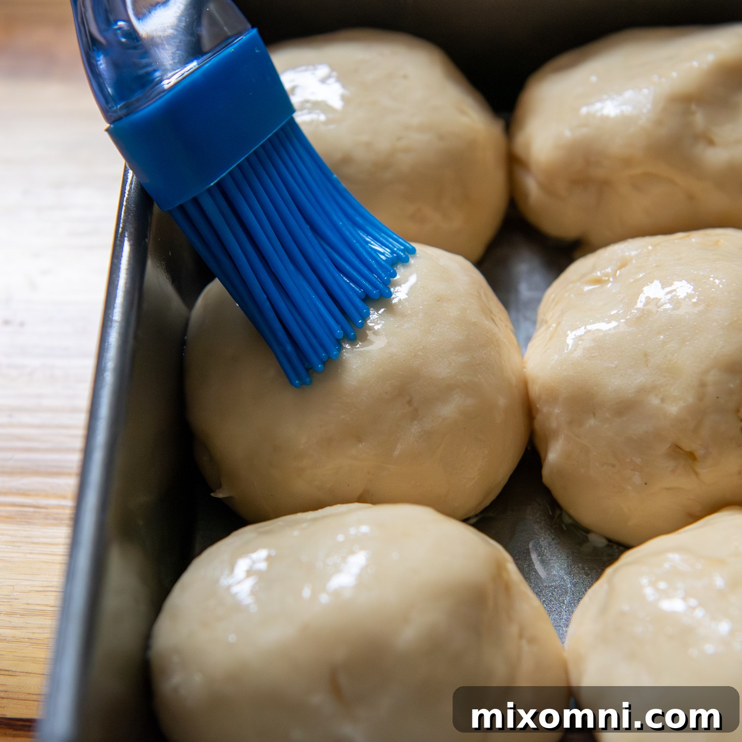A blue silicone brush gently painting melted butter onto the tops of risen, unbaked gluten-free Hawaiian rolls, enhancing their future golden crust.