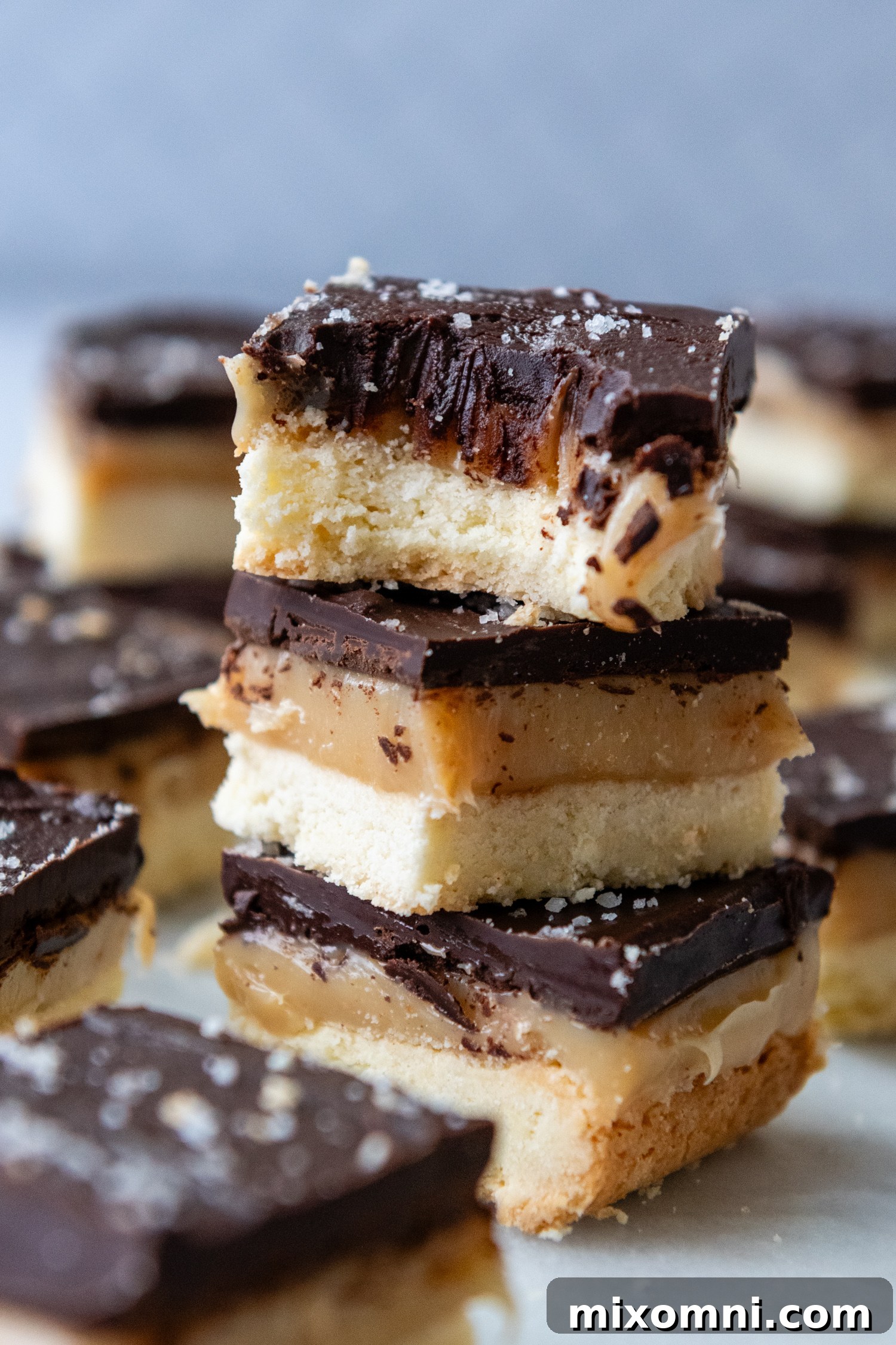 A stack of perfectly cut gluten free millionaire bars, with the top bar having a bite taken out, showing gooey caramel and crumbly shortbread.